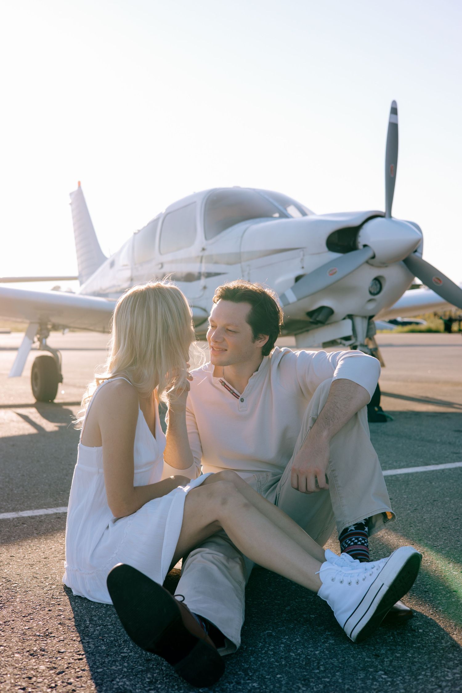 Happy couple sitting together on an airport tarmac in front of a white propeller plane in golden sunlight.