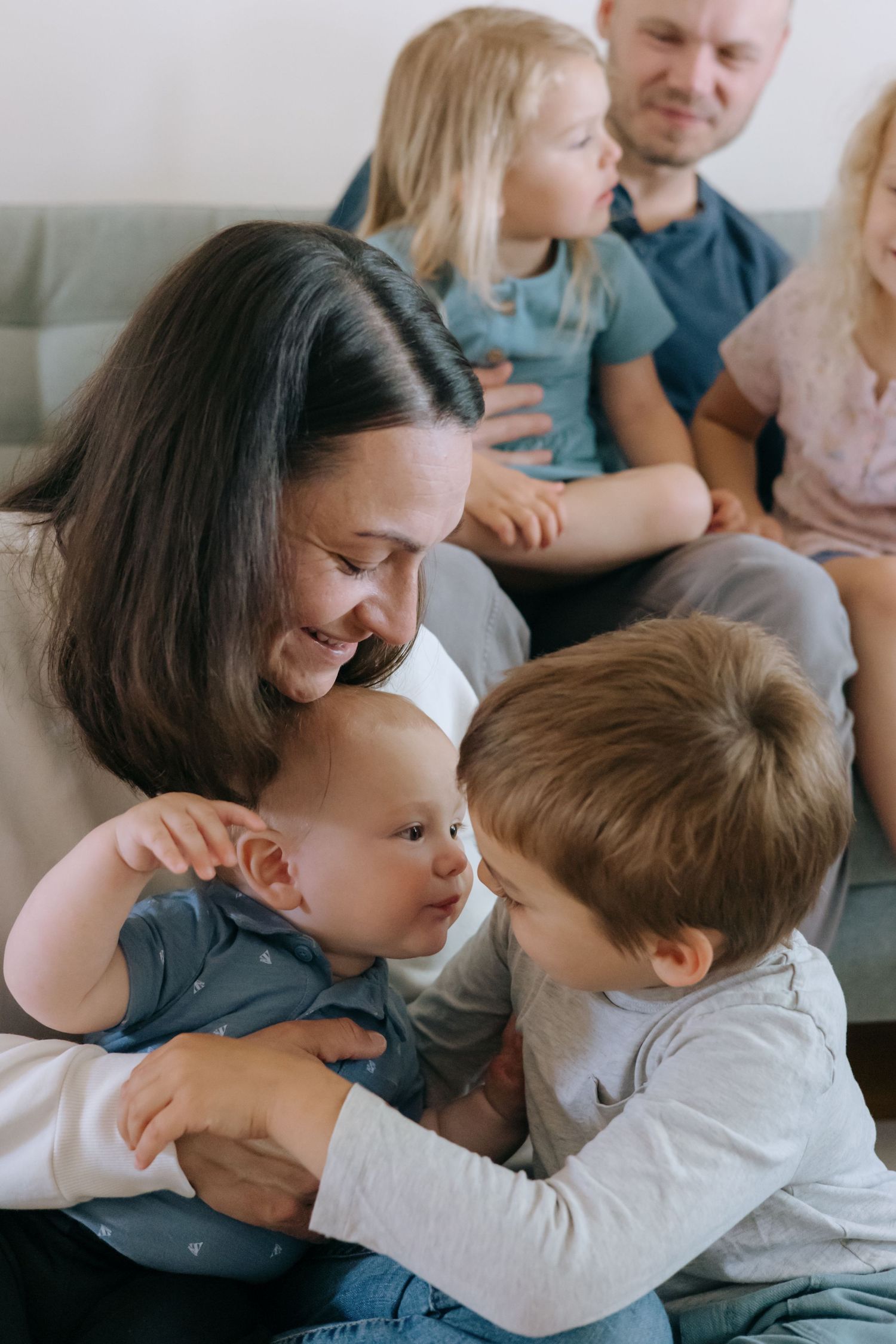 Happy family on couch with dark-haired mother smiling at baby while young son reaches out to touch infant.