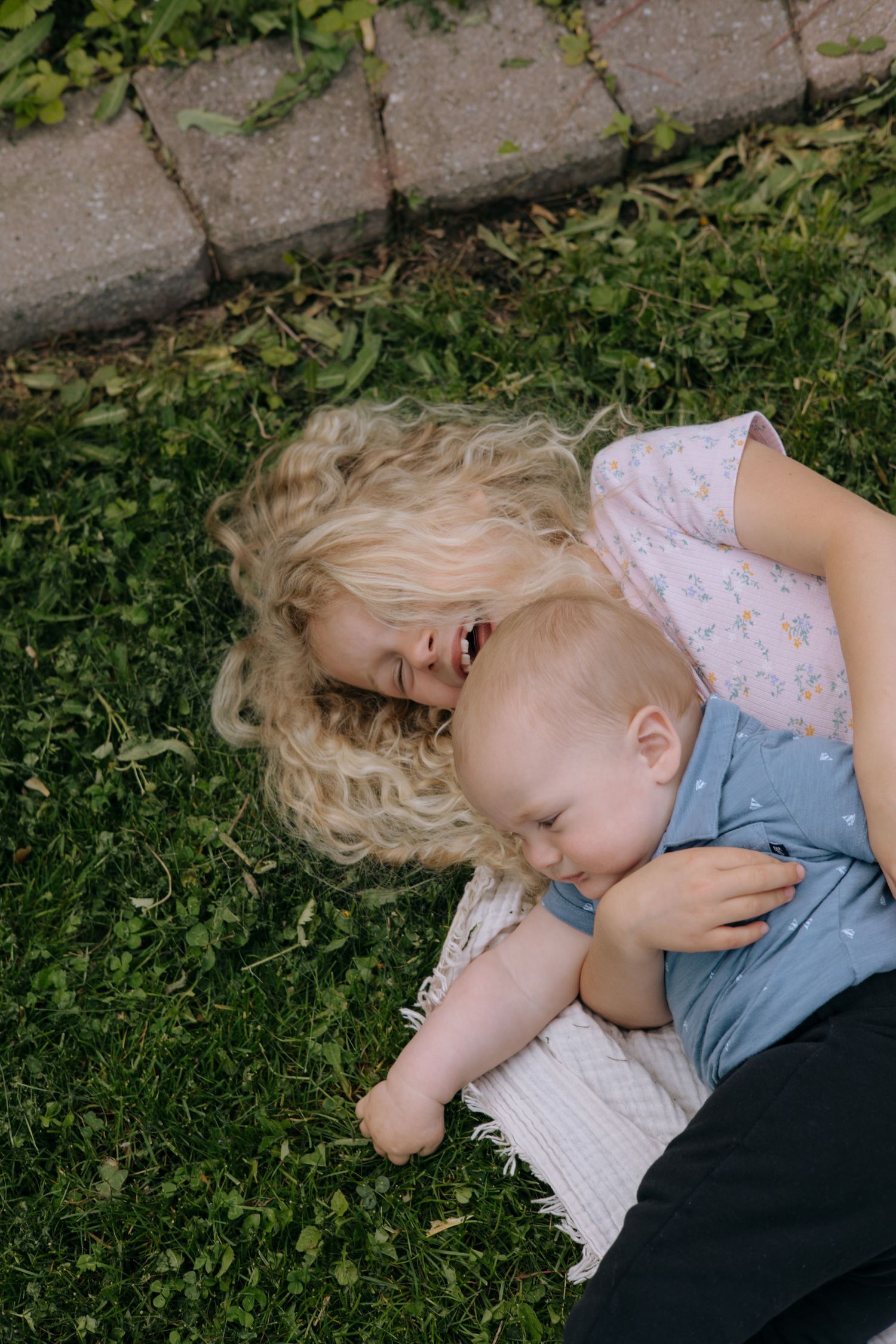 A young blonde girl lies on the grass outdoors, lovingly cuddling a baby on a blanket.