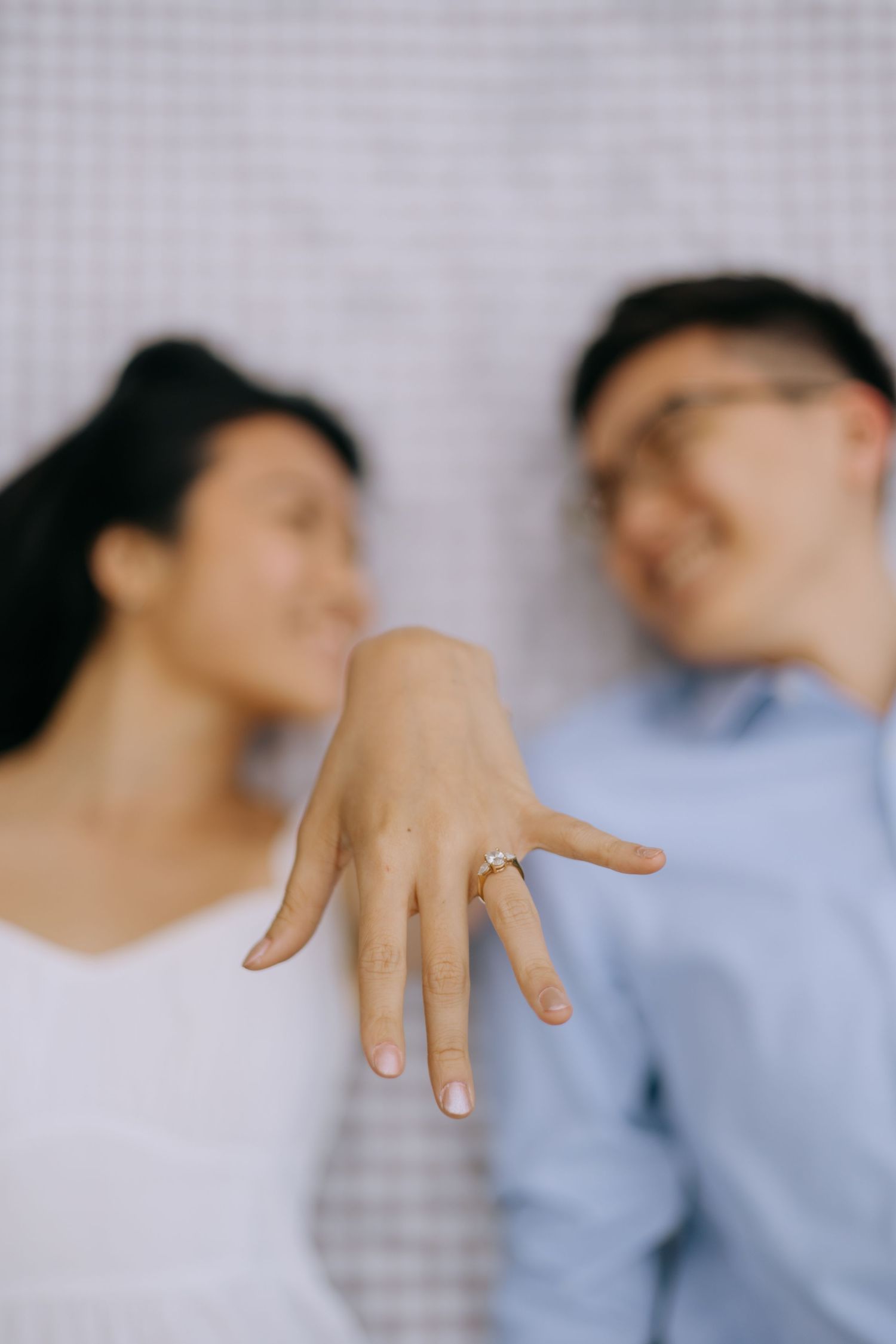 Engaged couple lying down smiling while woman displays diamond engagement ring toward camera.