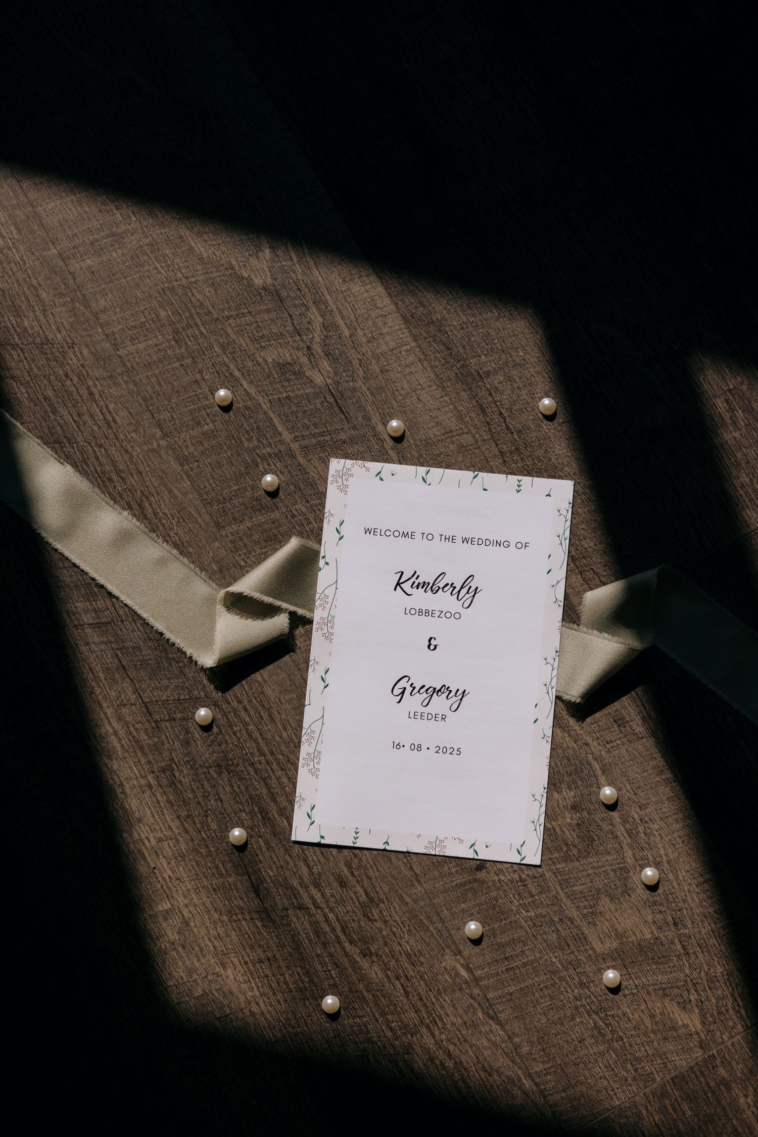 Elegant wedding program card displayed on a wooden surface with pearls and ribbon.