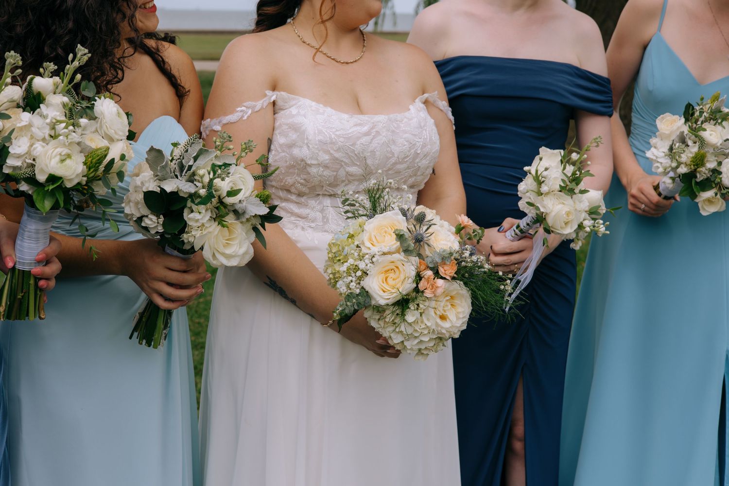 Bride and bridesmaids in blue dresses holding elegant white and green wedding bouquets outdoors.