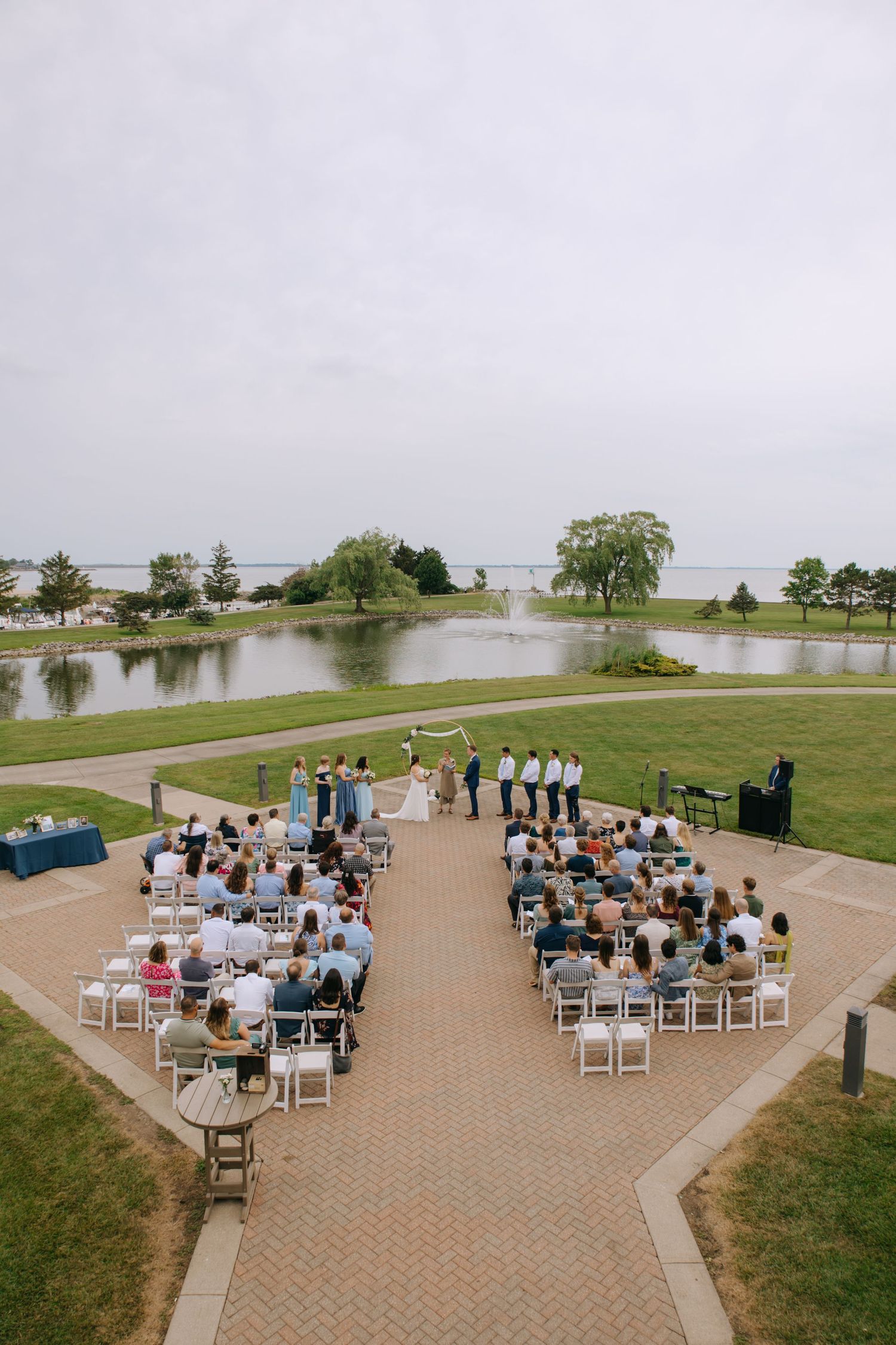 Aerial view of an outdoor waterfront wedding ceremony with guests seated on white chairs by a pond.