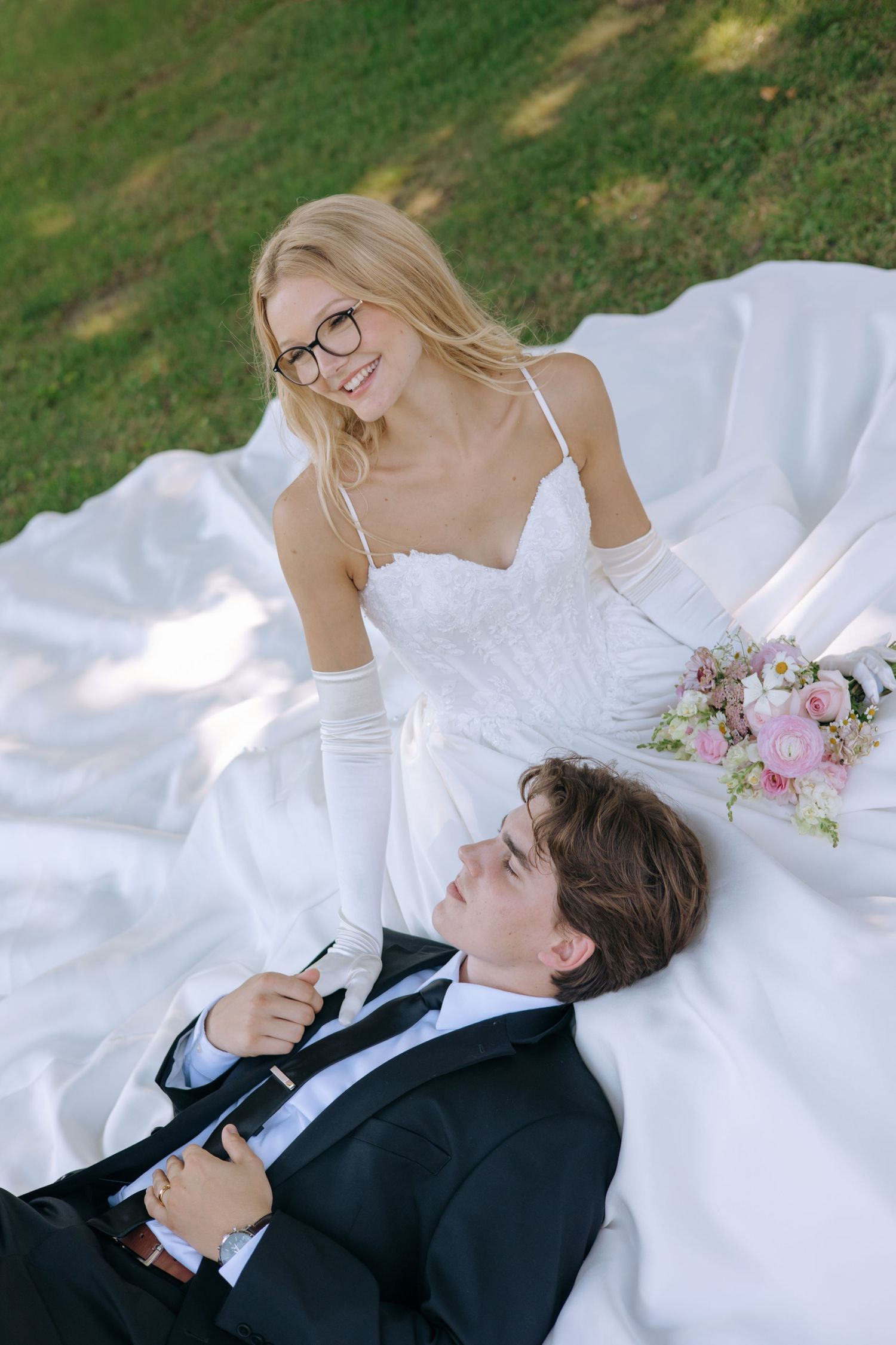 Bride in white gown and glasses sits smiling beside groom in black suit lying on wedding dress outdoors.