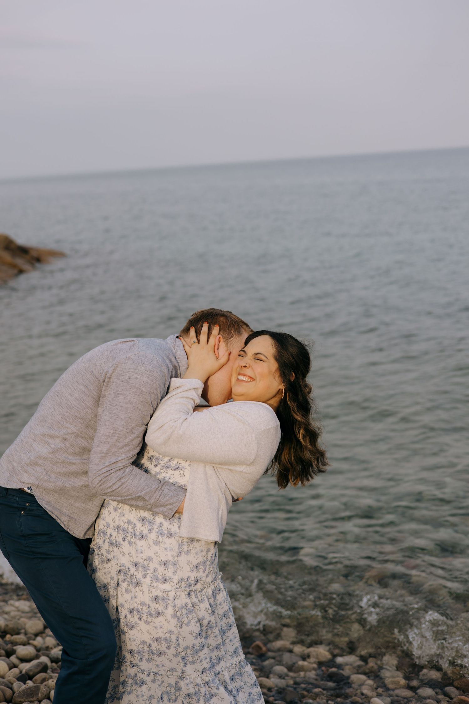 Man kissing woman on the cheek as she smiles joyfully beside a calm ocean shoreline.