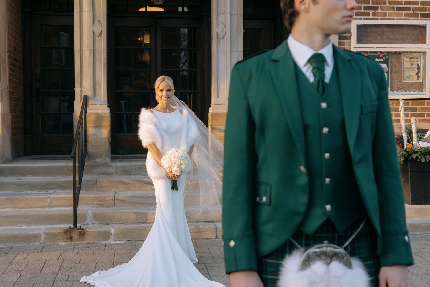 Bride in a fitted white gown with veil stands on church steps as groom in green Scottish kilt stands in foreground.