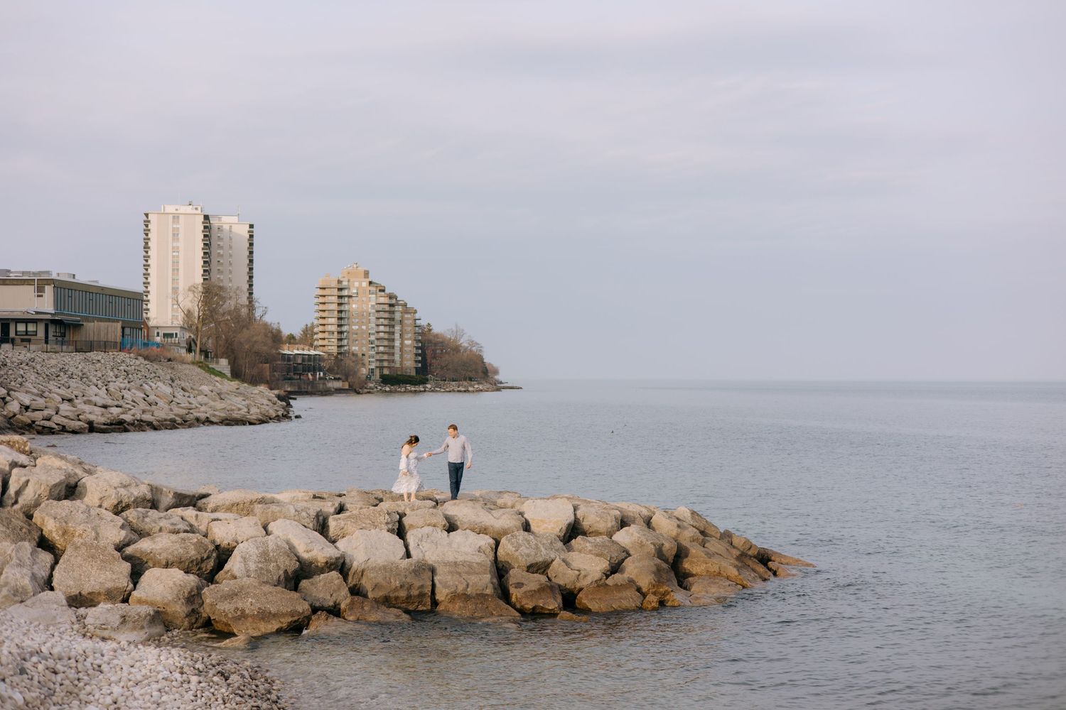 A couple stands on a rocky shoreline breakwater with city buildings and calm water in the background.