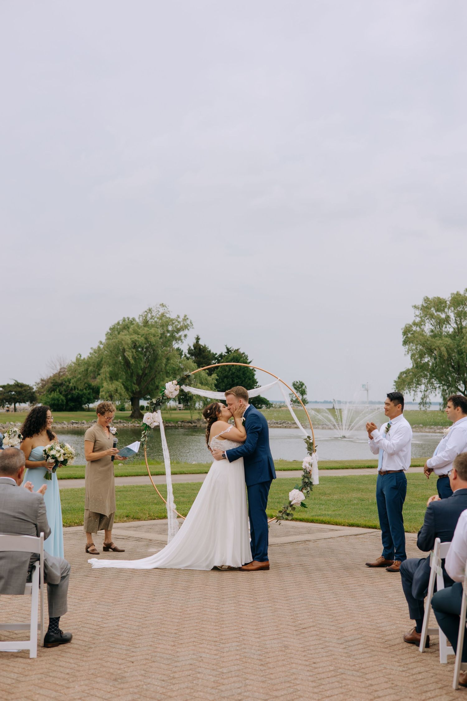 Bride and groom share first kiss at outdoor waterfront wedding ceremony with guests watching.