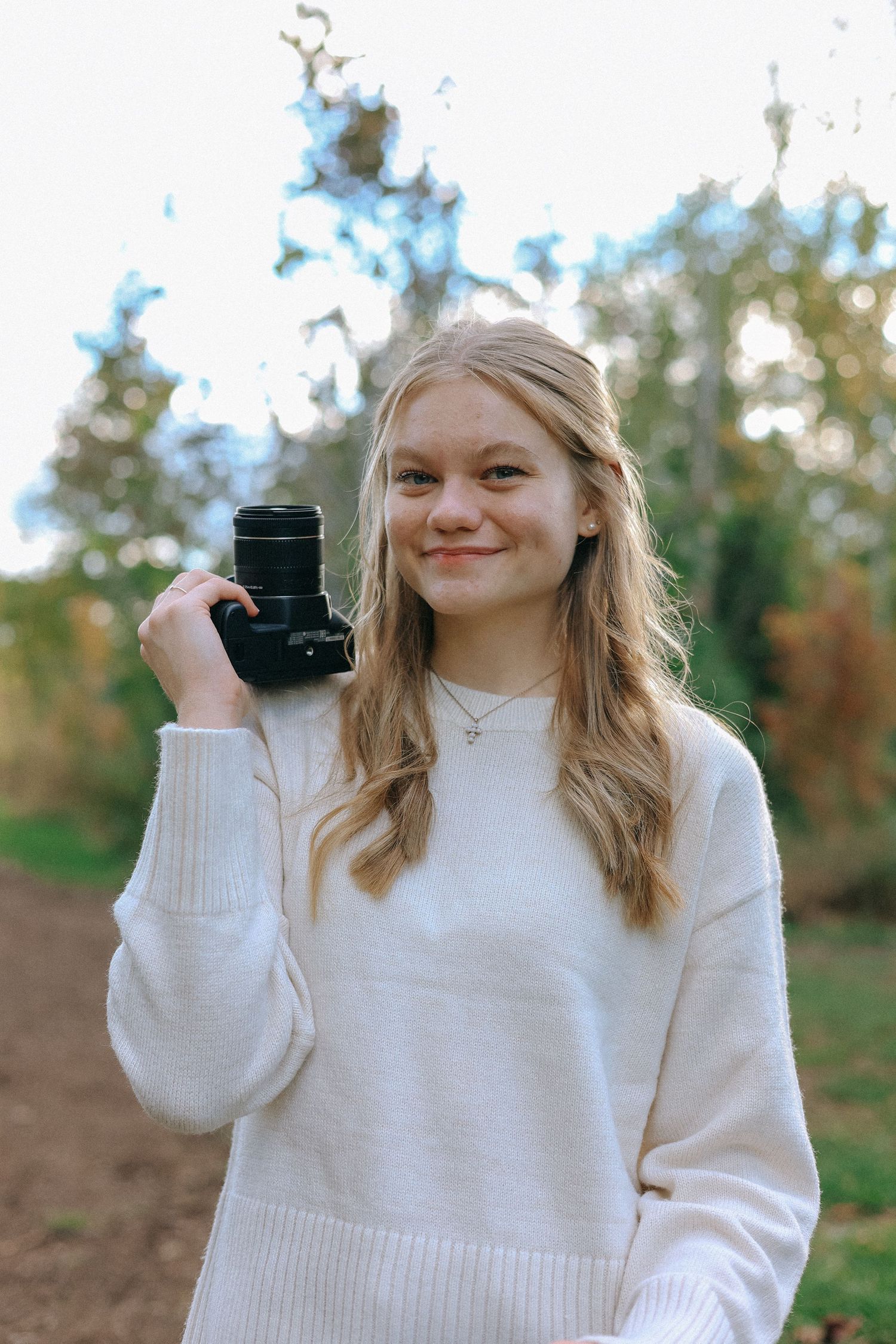 Smiling young blonde woman holding a camera outdoors in natural light wearing a white sweater.