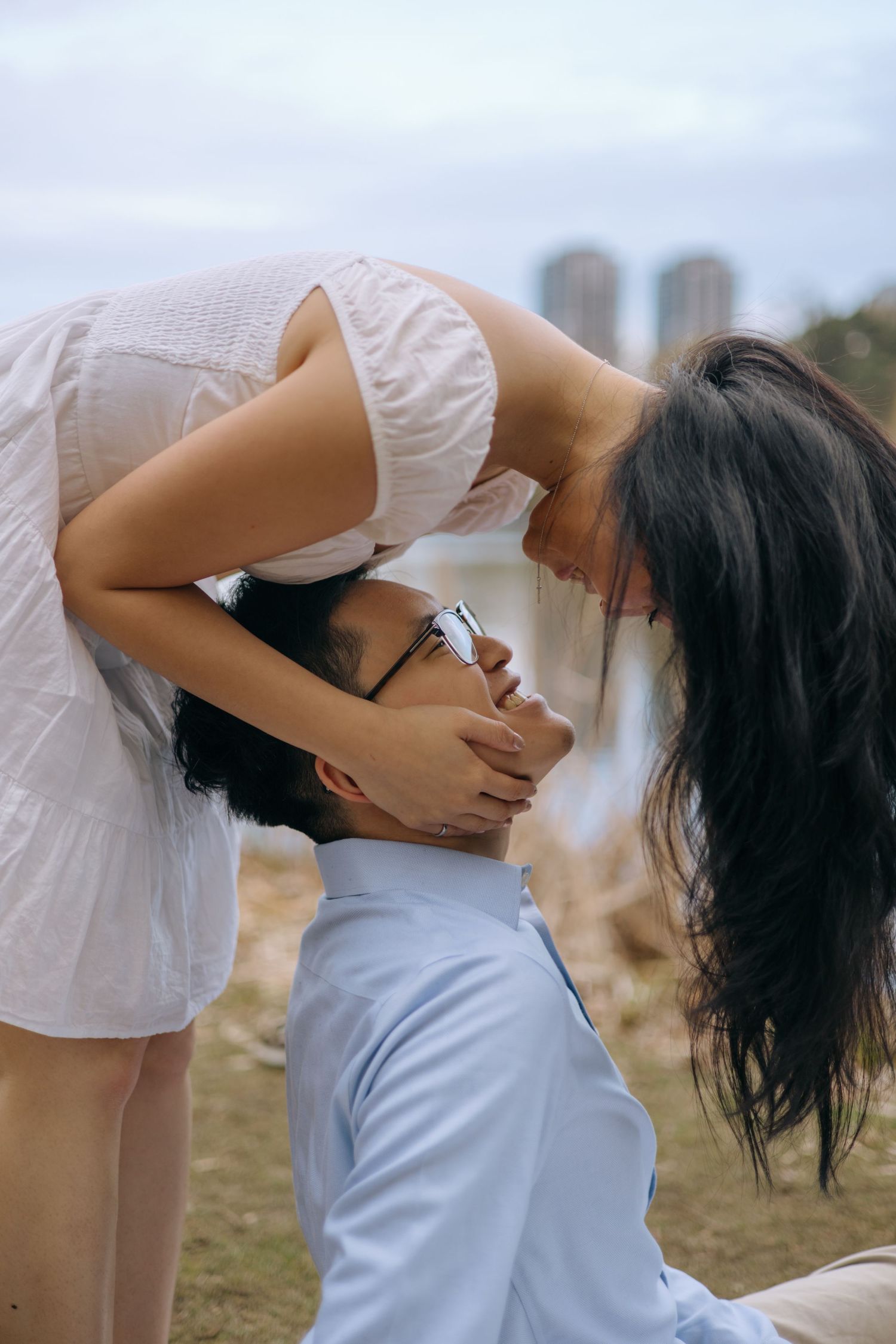 Romantic moment as woman in white dress leans down to kiss man sitting on ground outdoors.
