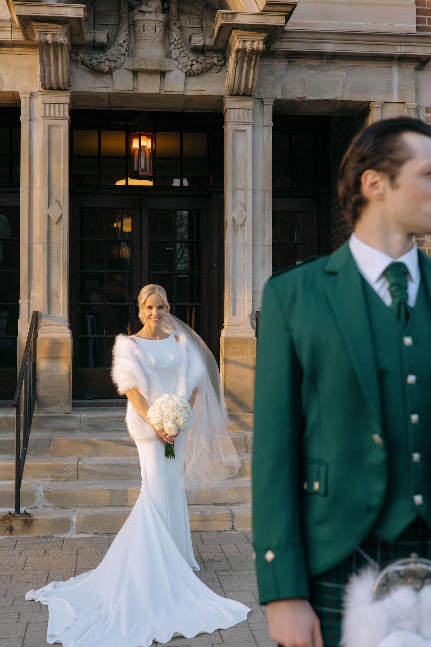 Elegant blonde bride in fitted white gown with fur wrap holds bouquet on steps of a grand stone building.