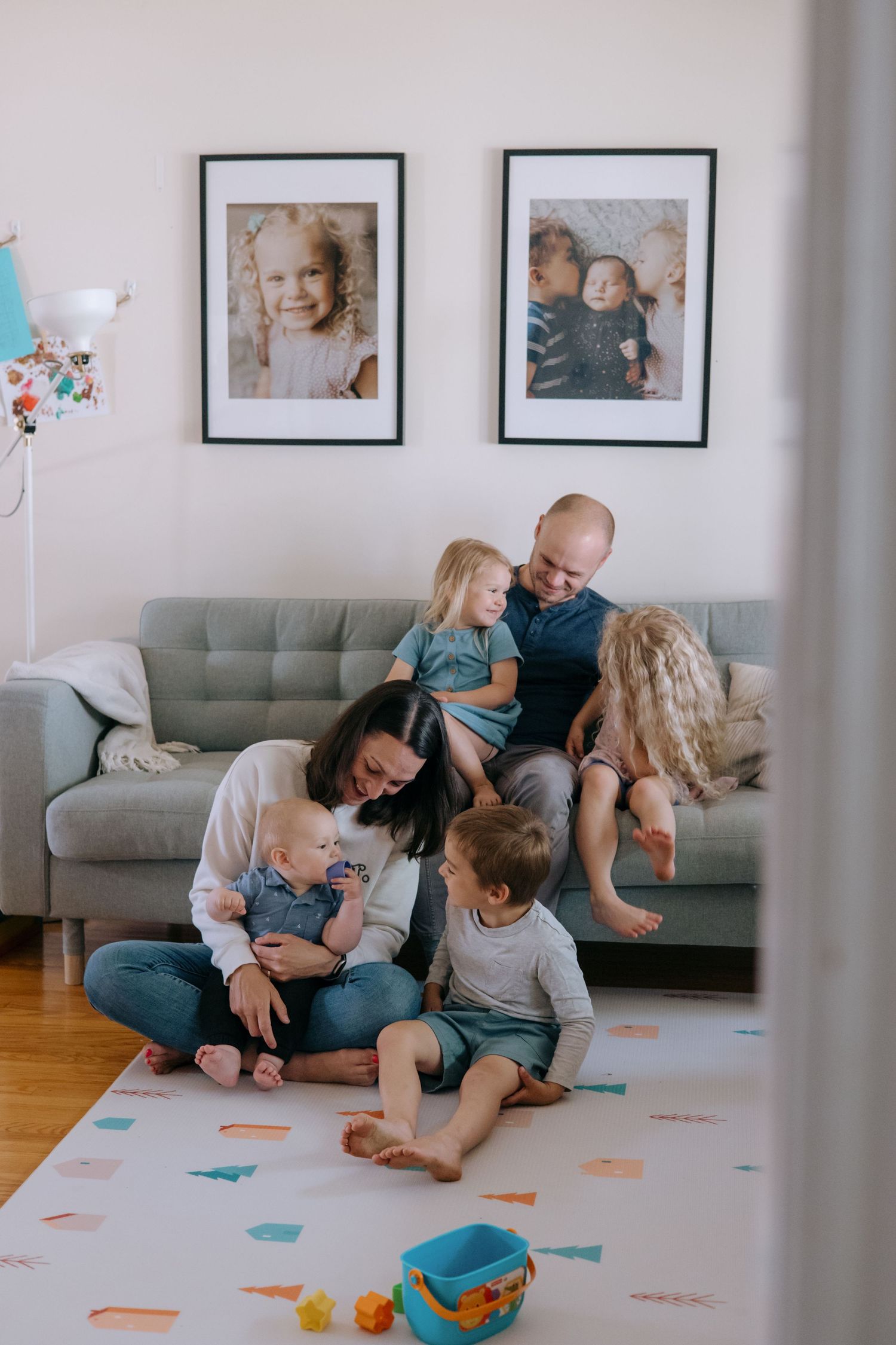 Young family of six relaxing together on living room floor with framed children photos displayed on wall behind them.