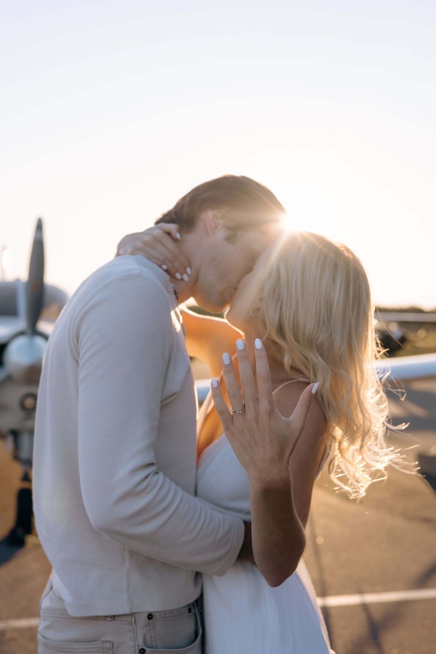 Couple sharing a romantic kiss in front of a small aircraft during a stunning golden sunset.