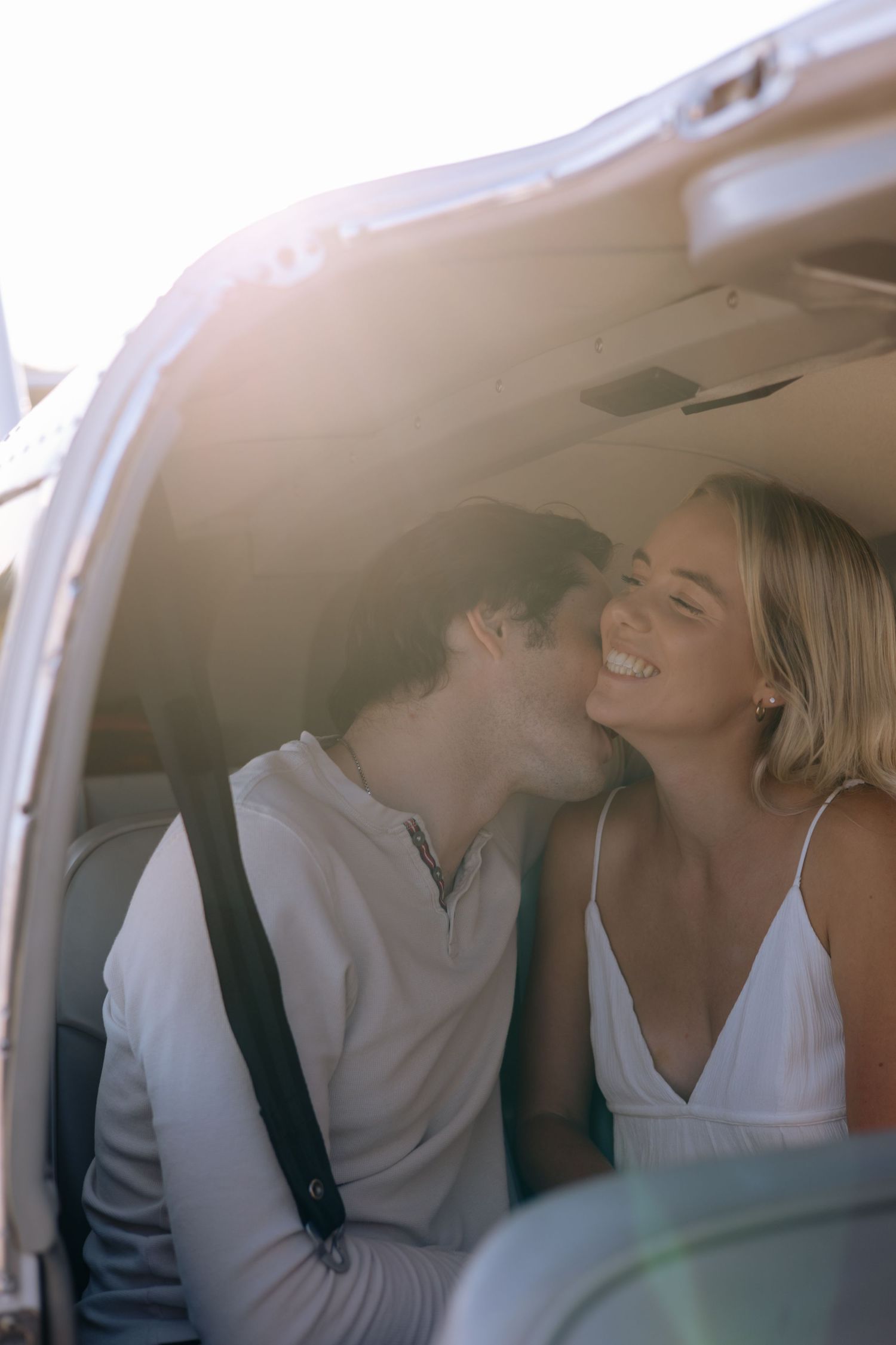 Joyful woman smiles as man kisses her cheek inside a small aircraft bathed in warm golden sunlight.