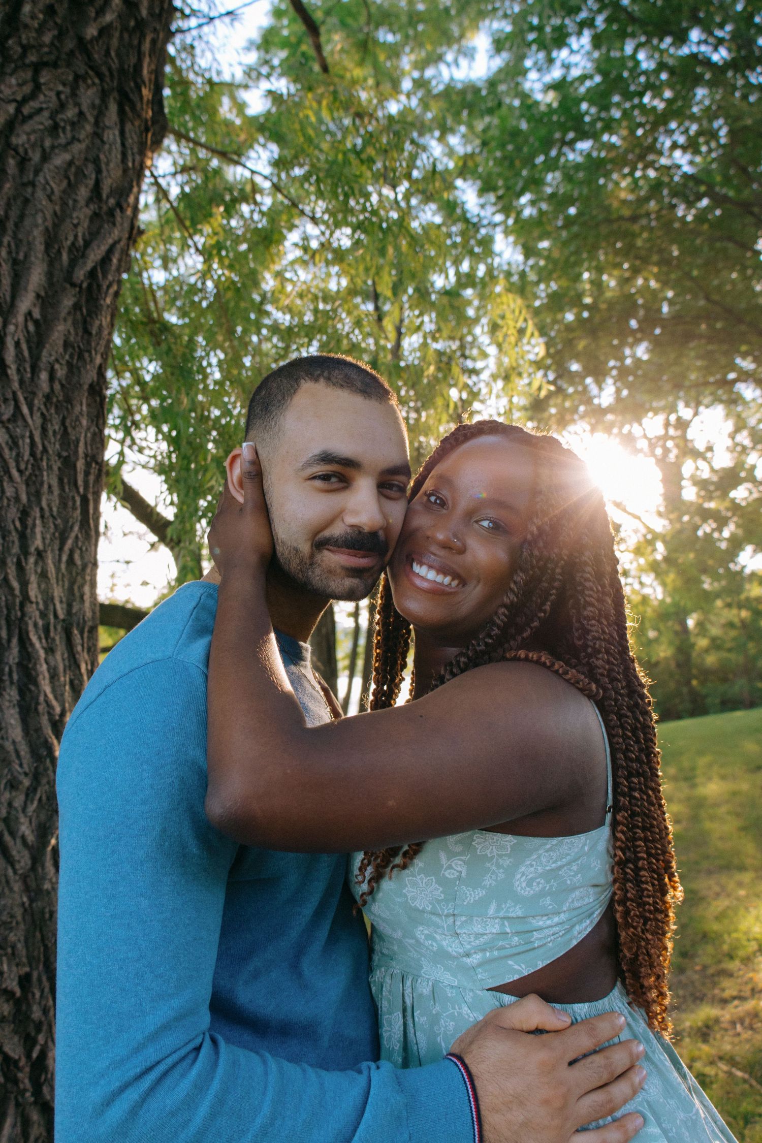 Happy interracial couple embracing and smiling by a tree with golden sunset light behind them.