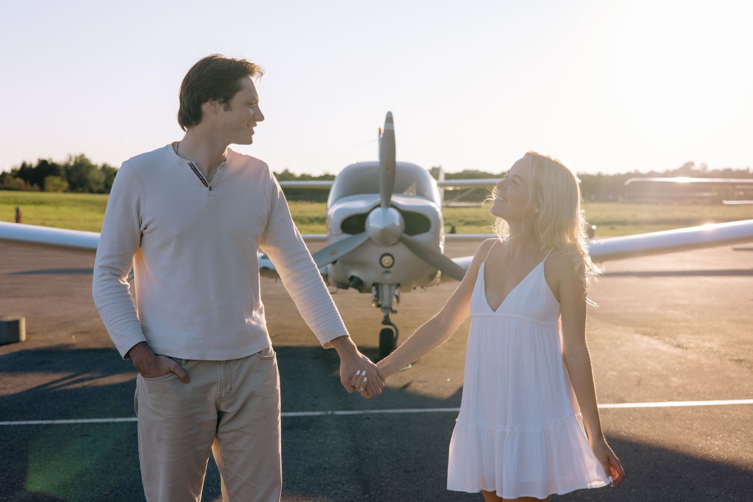 Smiling couple holding hands on a sunny airfield with a small private plane in the background.