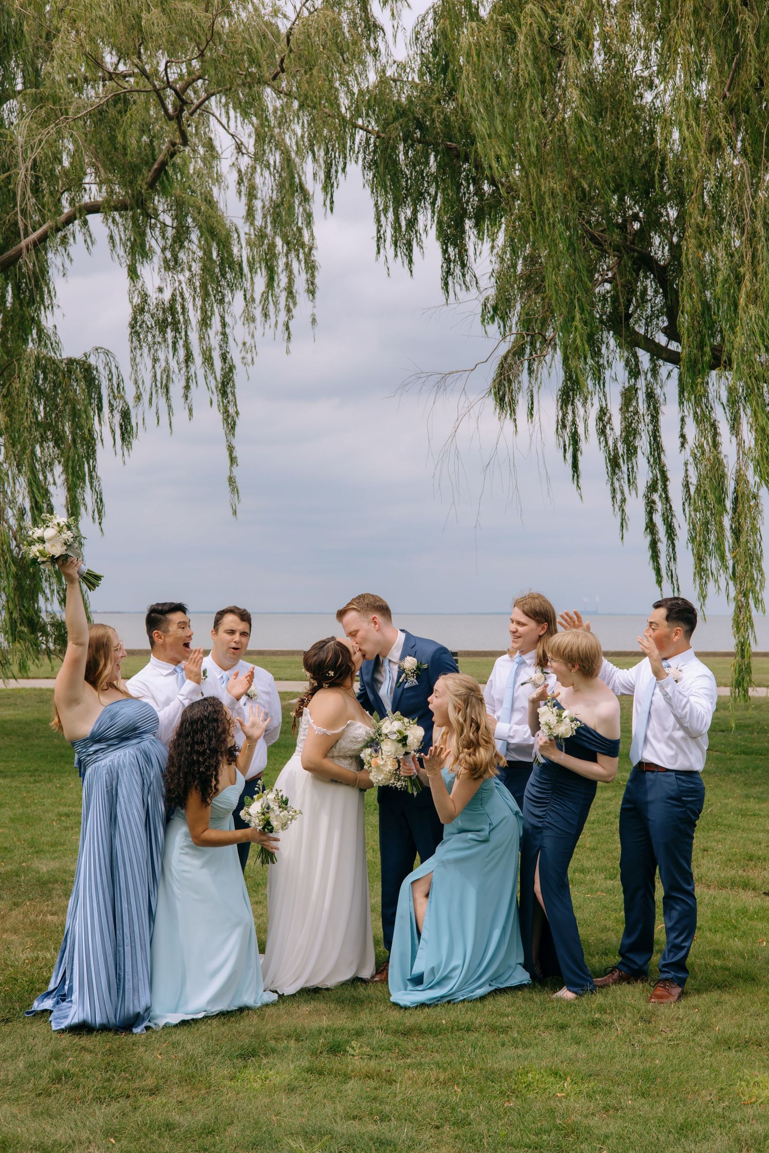 Wedding party celebrating under weeping willow tree, bridesmaids in blue dresses with bride and groom kissing outdoors.