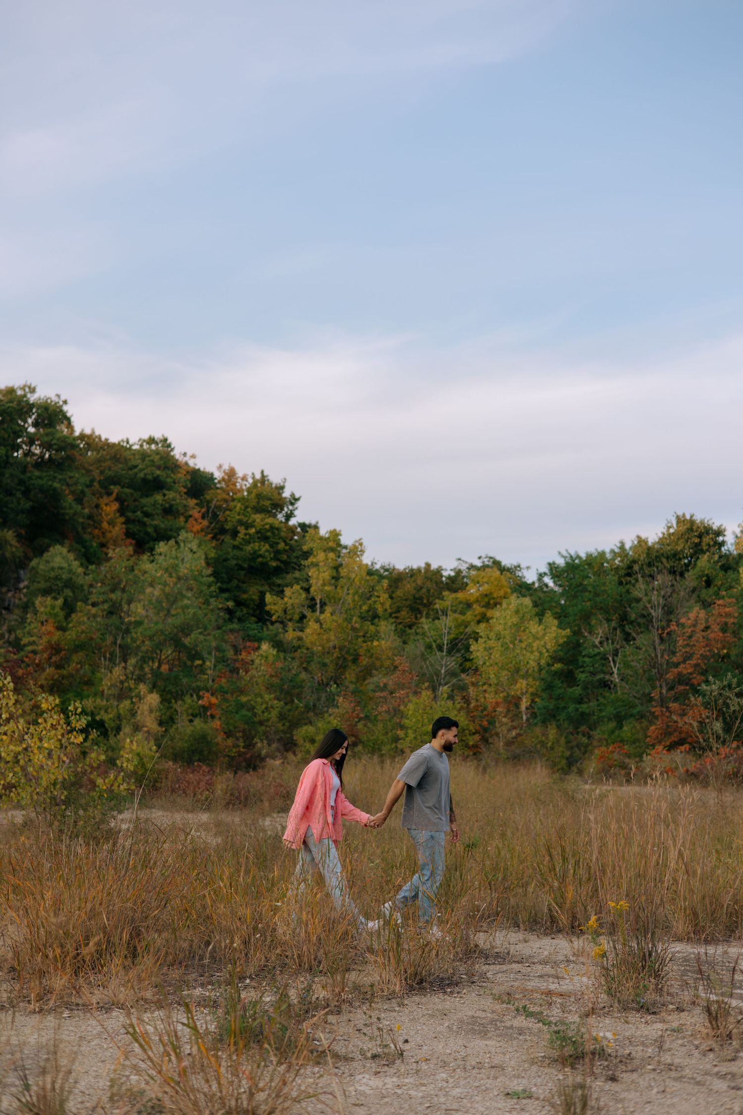 Couple holding hands while walking through an open field with colorful autumn trees in background.