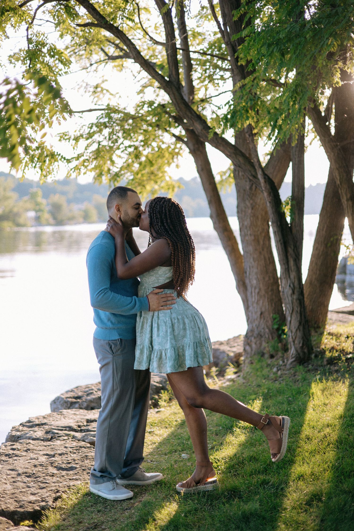 Romantic interracial couple sharing a kiss near a sunlit lake surrounded by green trees.