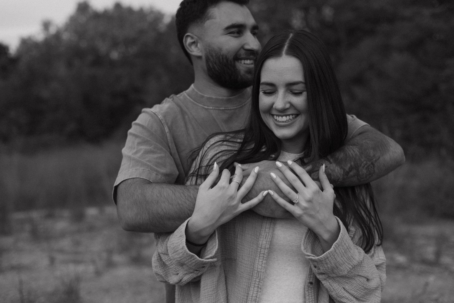 Black and white photo of man embracing woman from behind, both smiling in an outdoor setting.