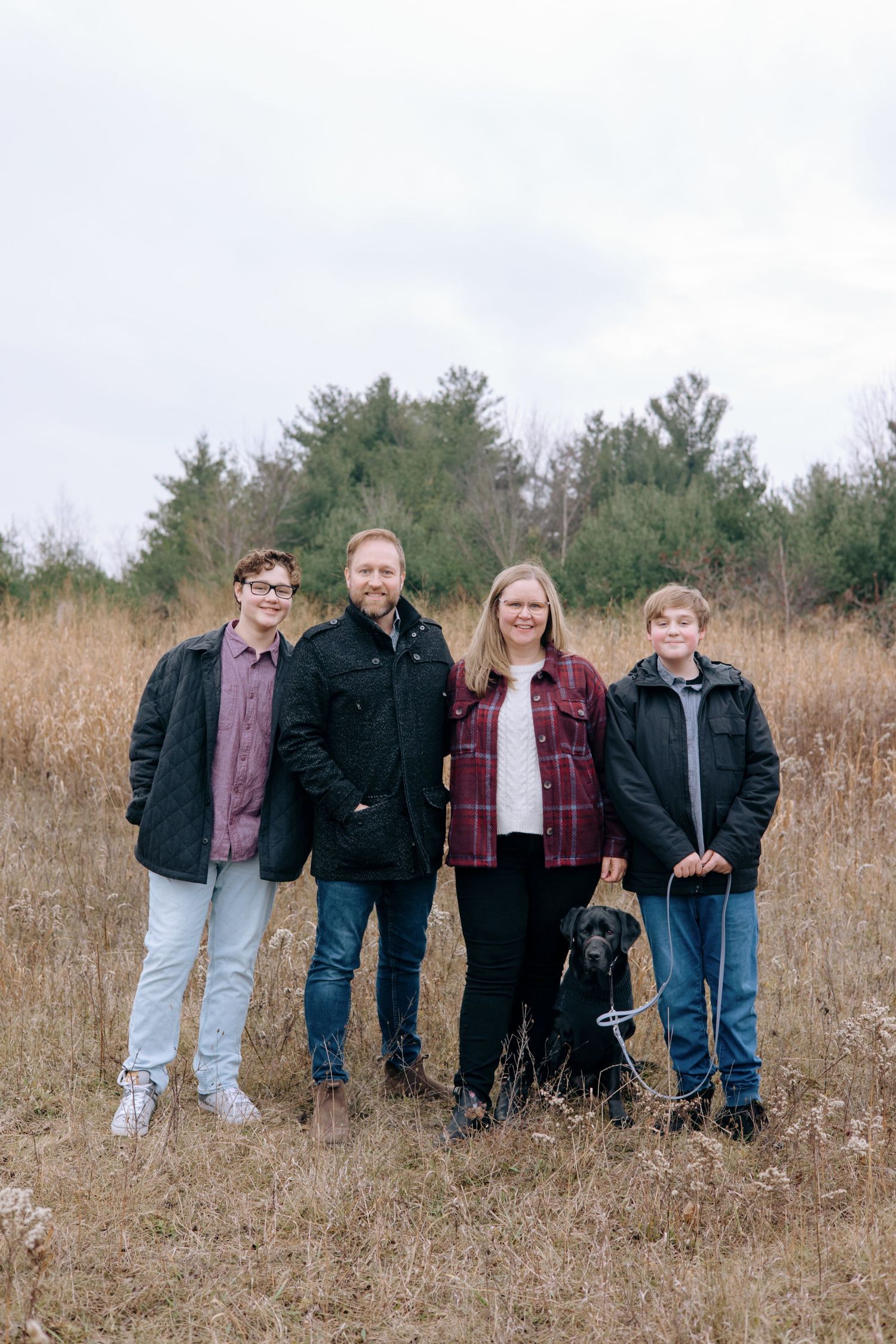 Family of four posing with black Labrador in dry autumn field surrounded by trees on overcast day.