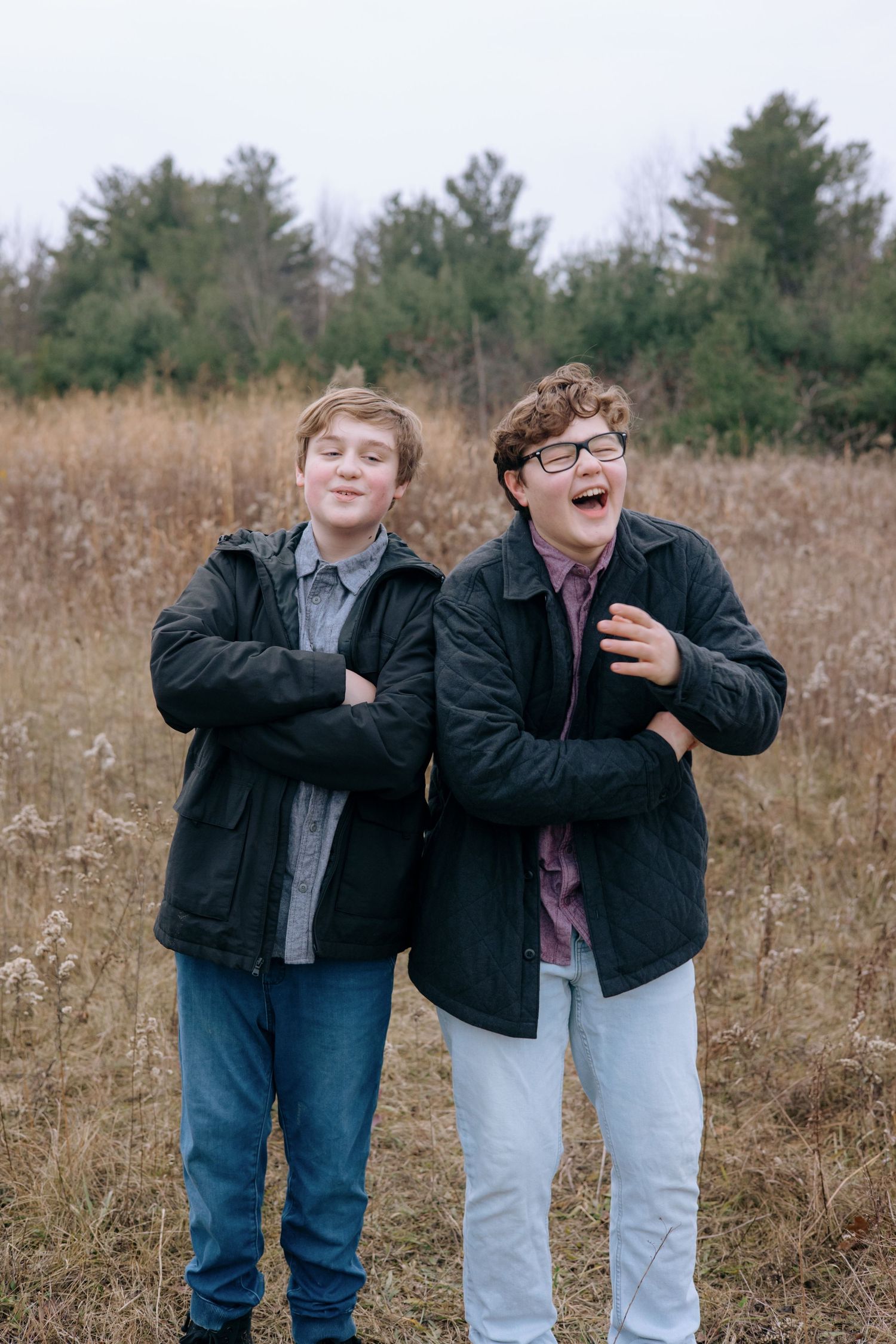 Two teenage brothers laughing together in autumn field wearing black jackets, one with glasses, in candid portrait.
