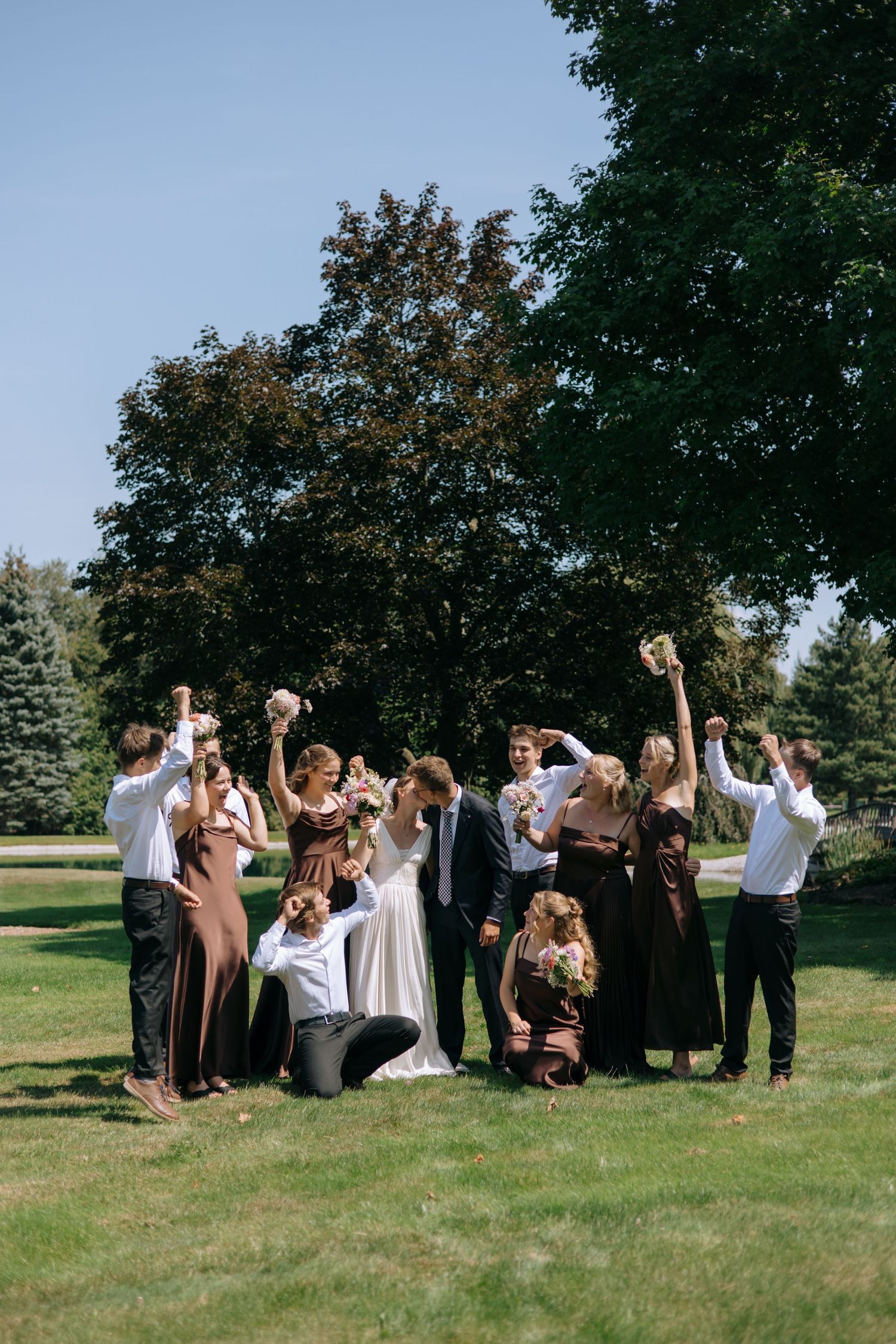 Wedding party celebrating outdoors on green lawn with bride, groom, bridesmaids in mauve dresses, and groomsmen cheering under trees.