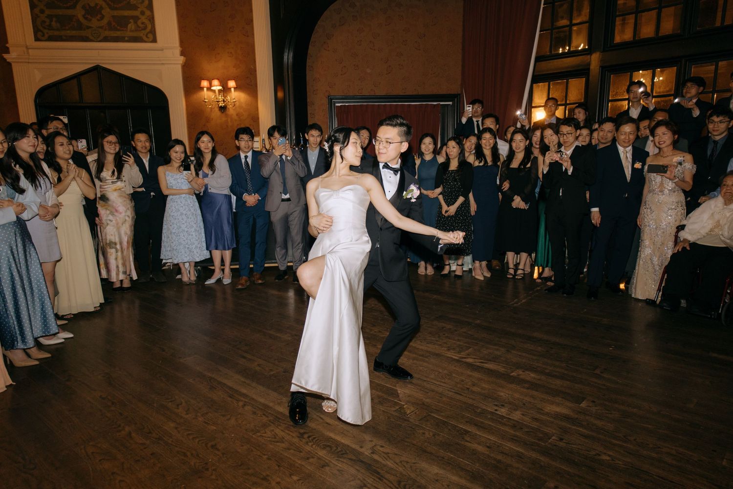 Bride and groom share first dance at elegant wedding reception with guests watching on wooden dance floor.