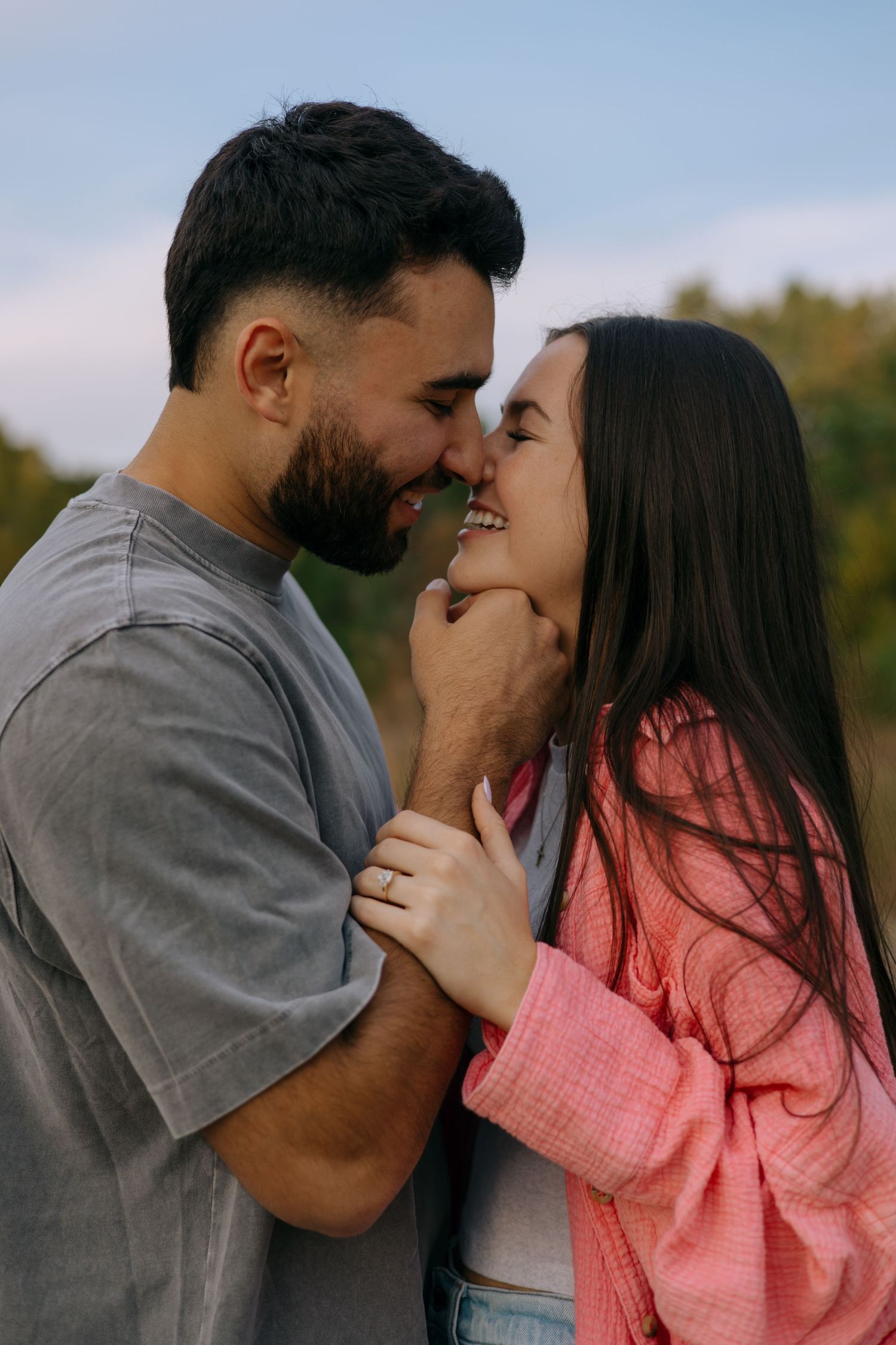 Happy couple sharing a romantic moment outdoors, smiling and touching foreheads against a blue sky.