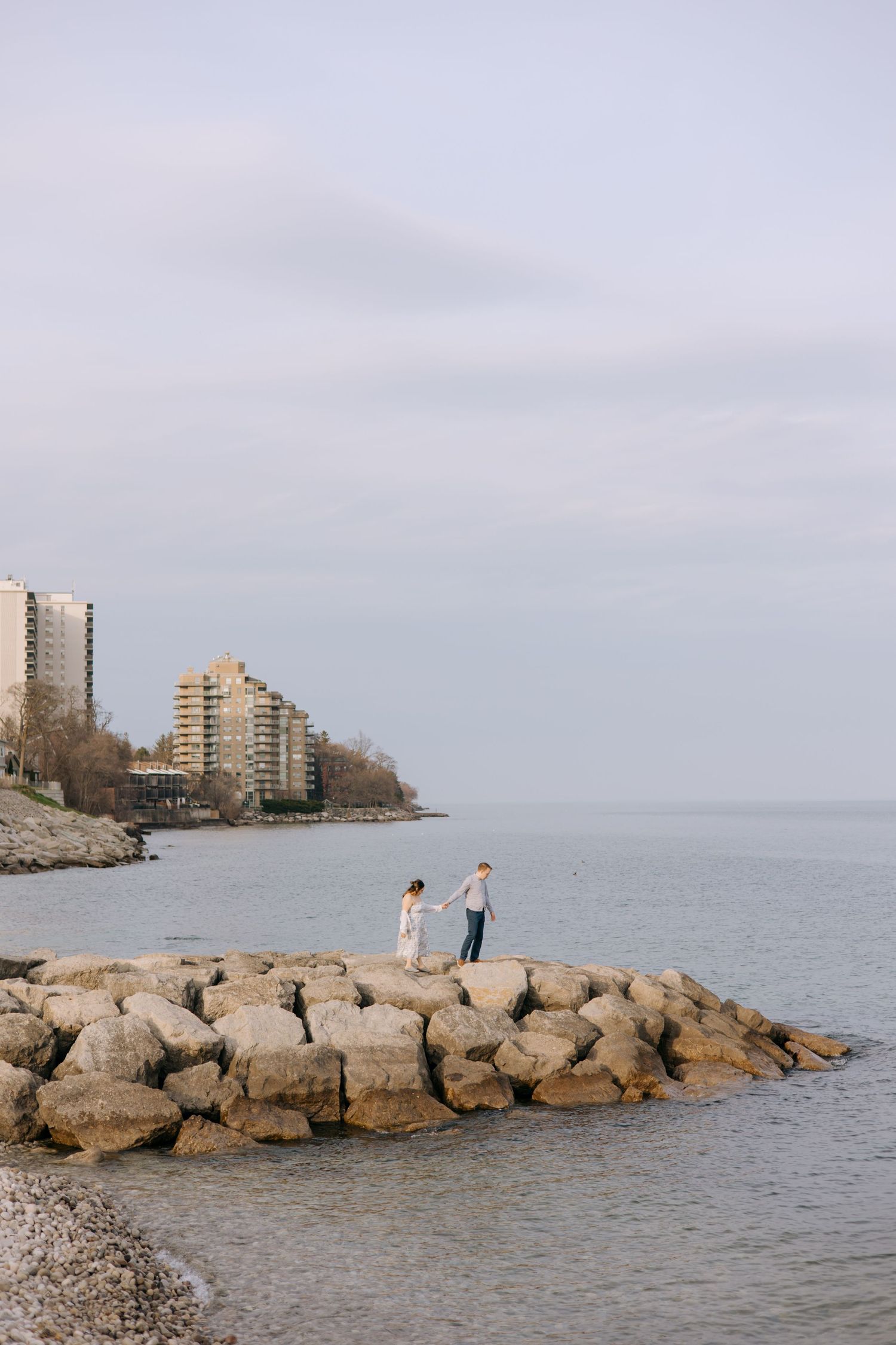 Couple holding hands on a rocky lakeside jetty with city buildings visible in the background.
