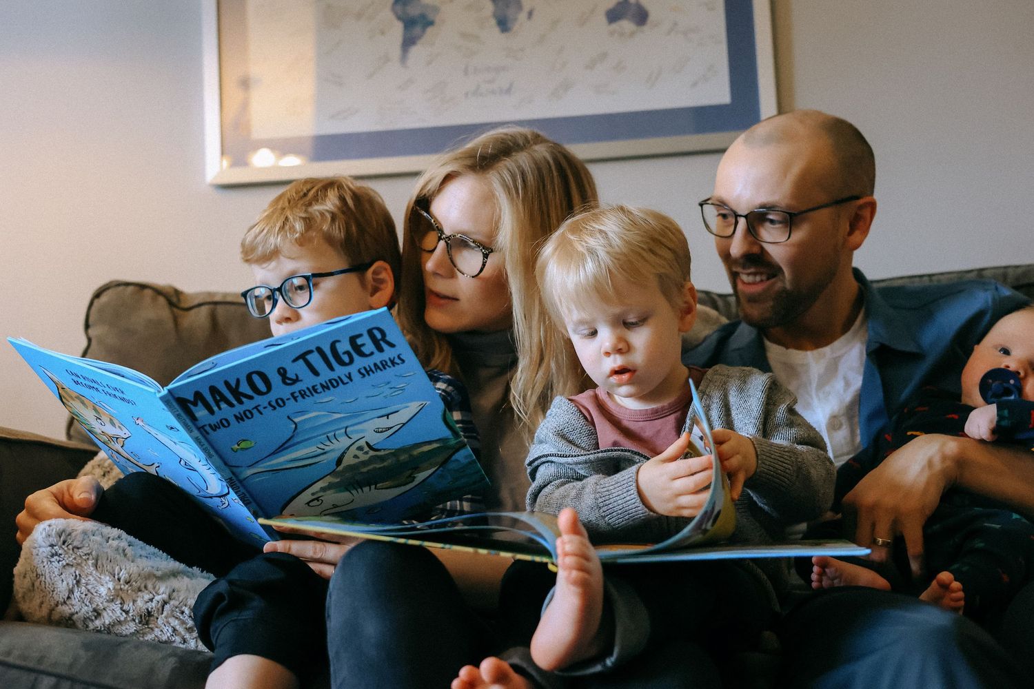 Family of four sitting together on a couch reading a colorful children's book in a cozy living room.