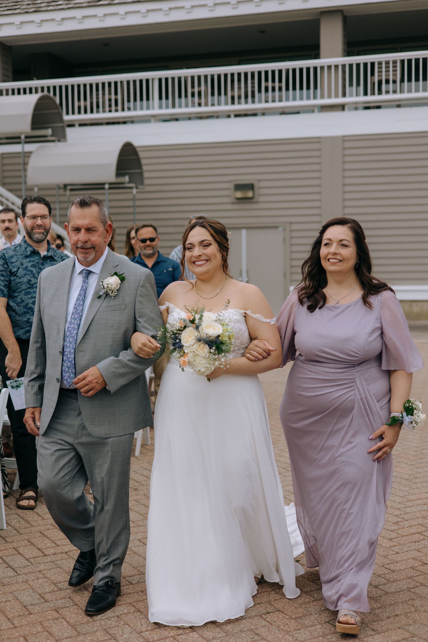 Smiling bride walks down the aisle escorted by her parents at an outdoor summer wedding ceremony.