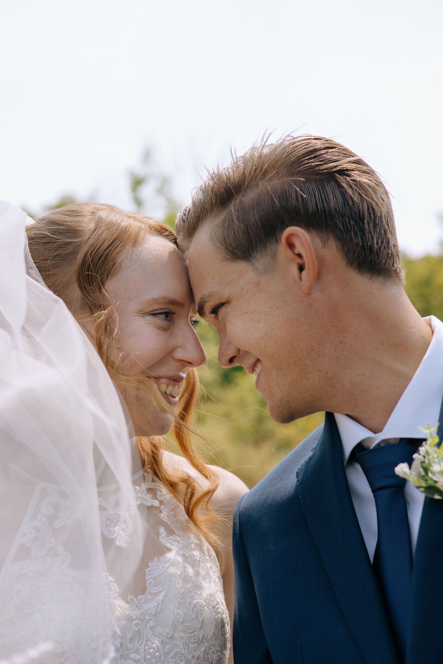 Bride and groom touching foreheads and smiling at each other outdoors on a sunny wedding day.