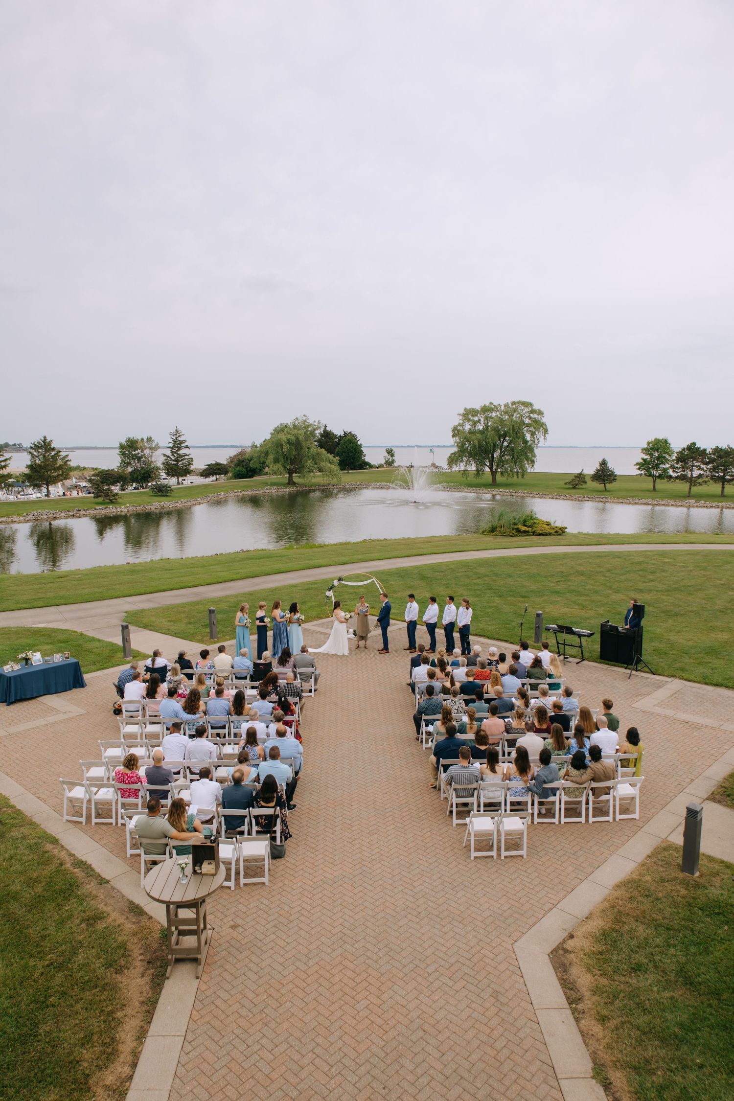 Aerial view of outdoor wedding ceremony with guests seated in white chairs facing altar near scenic pond.
