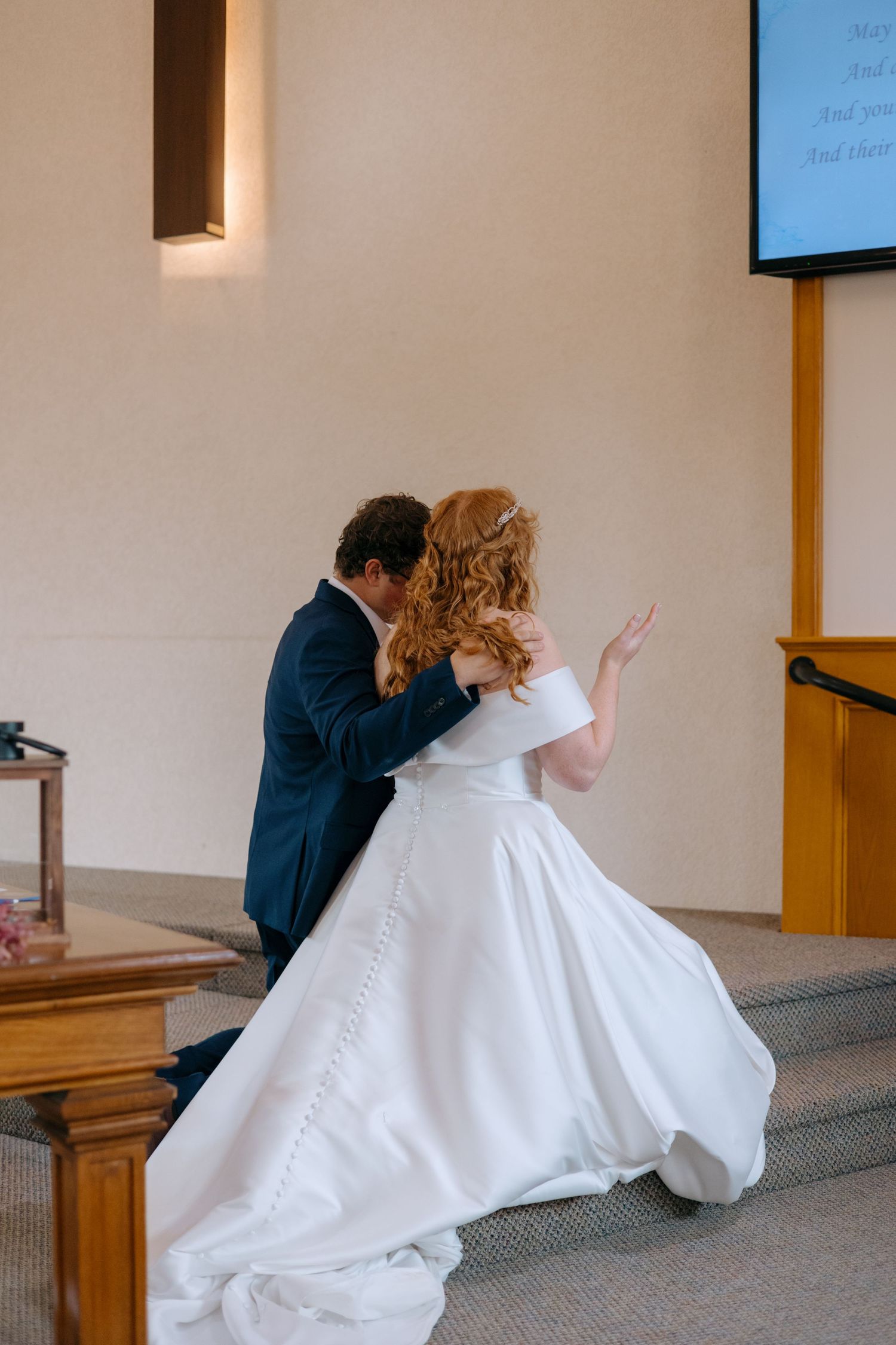 Bride in white off-shoulder gown and groom in navy suit embracing at church altar during ceremony.