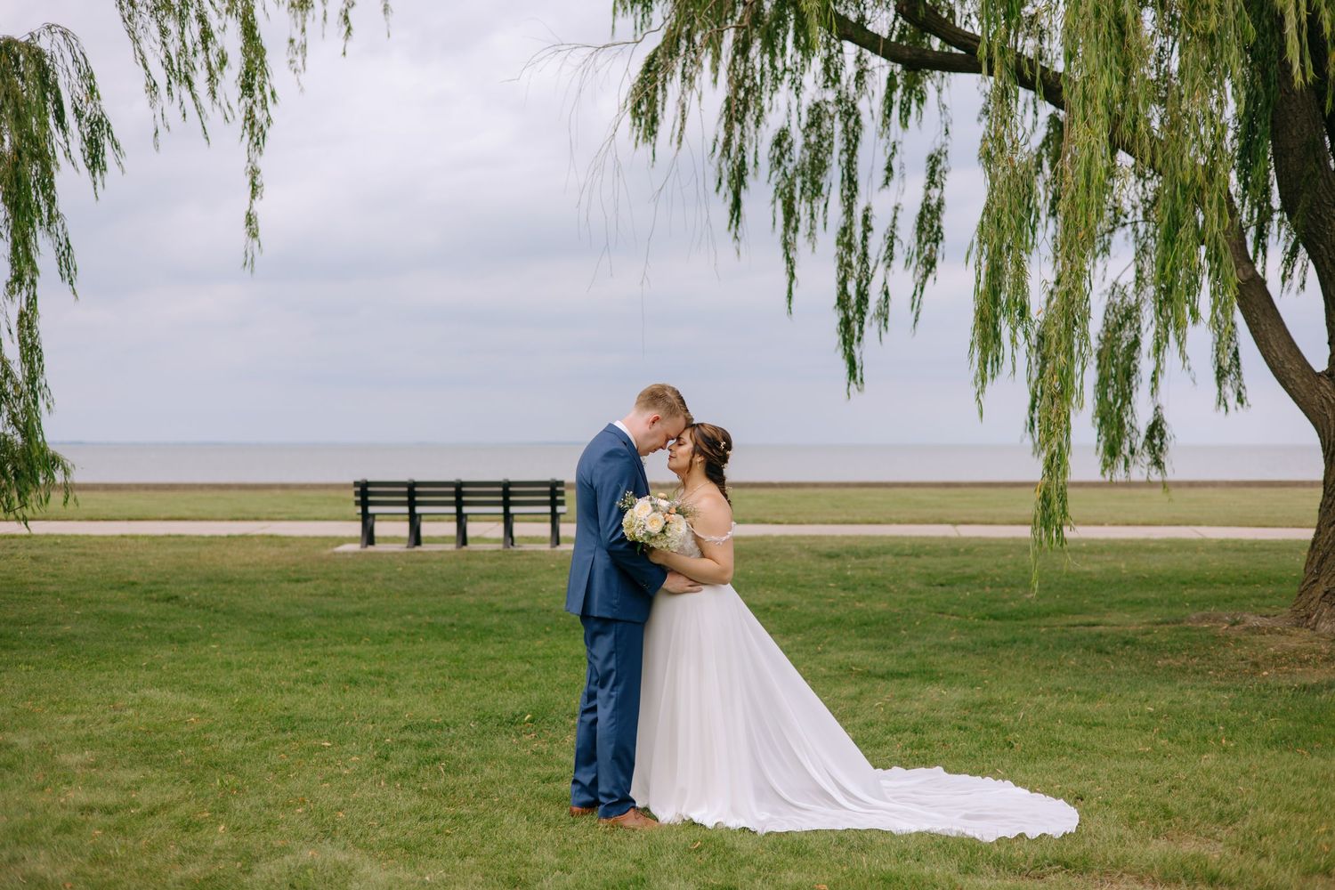 Bride and groom kiss romantically on a green lawn near a willow tree with a serene lakeside backdrop.