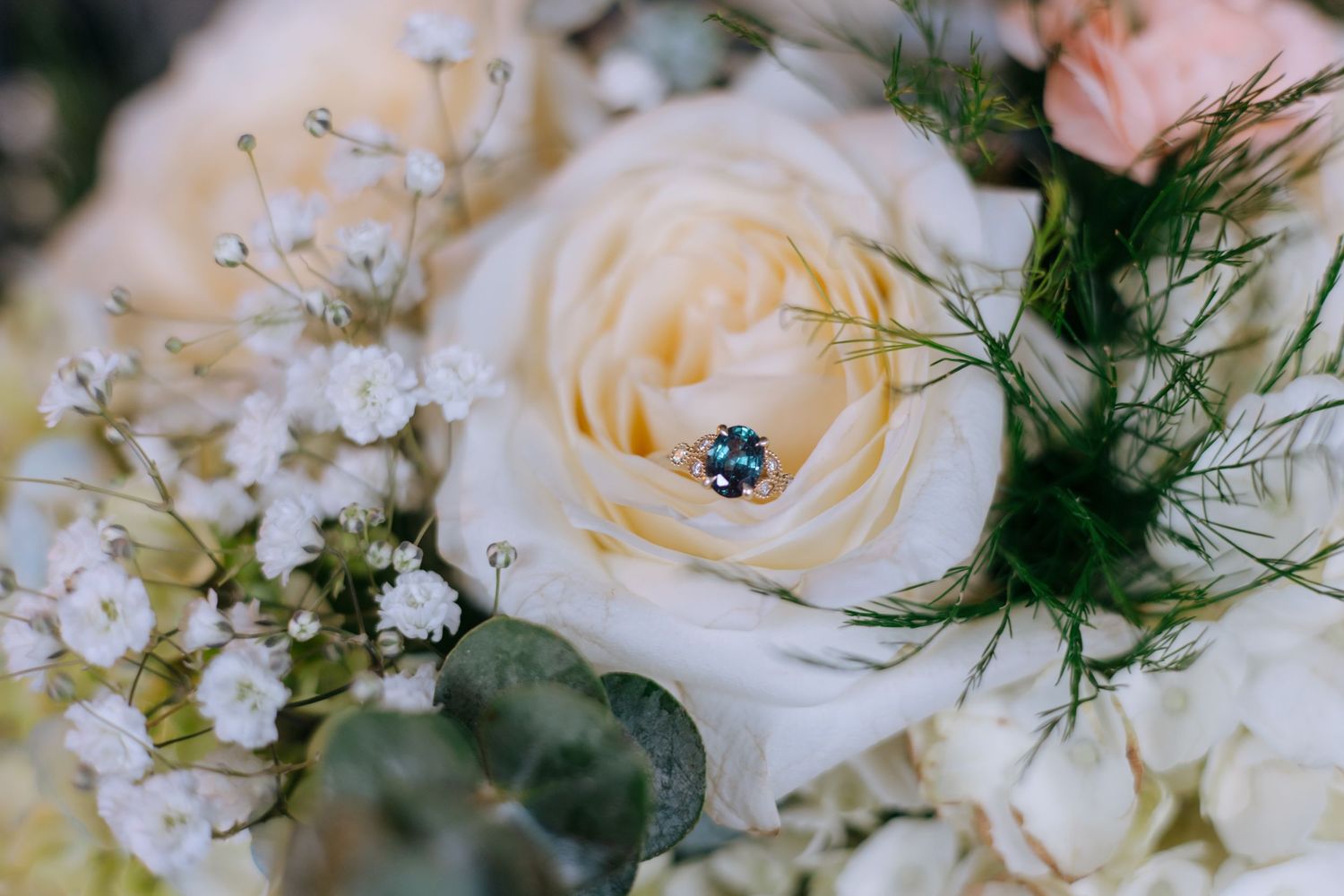 Elegant blue gemstone engagement ring resting on a white garden rose in a bridal bouquet.