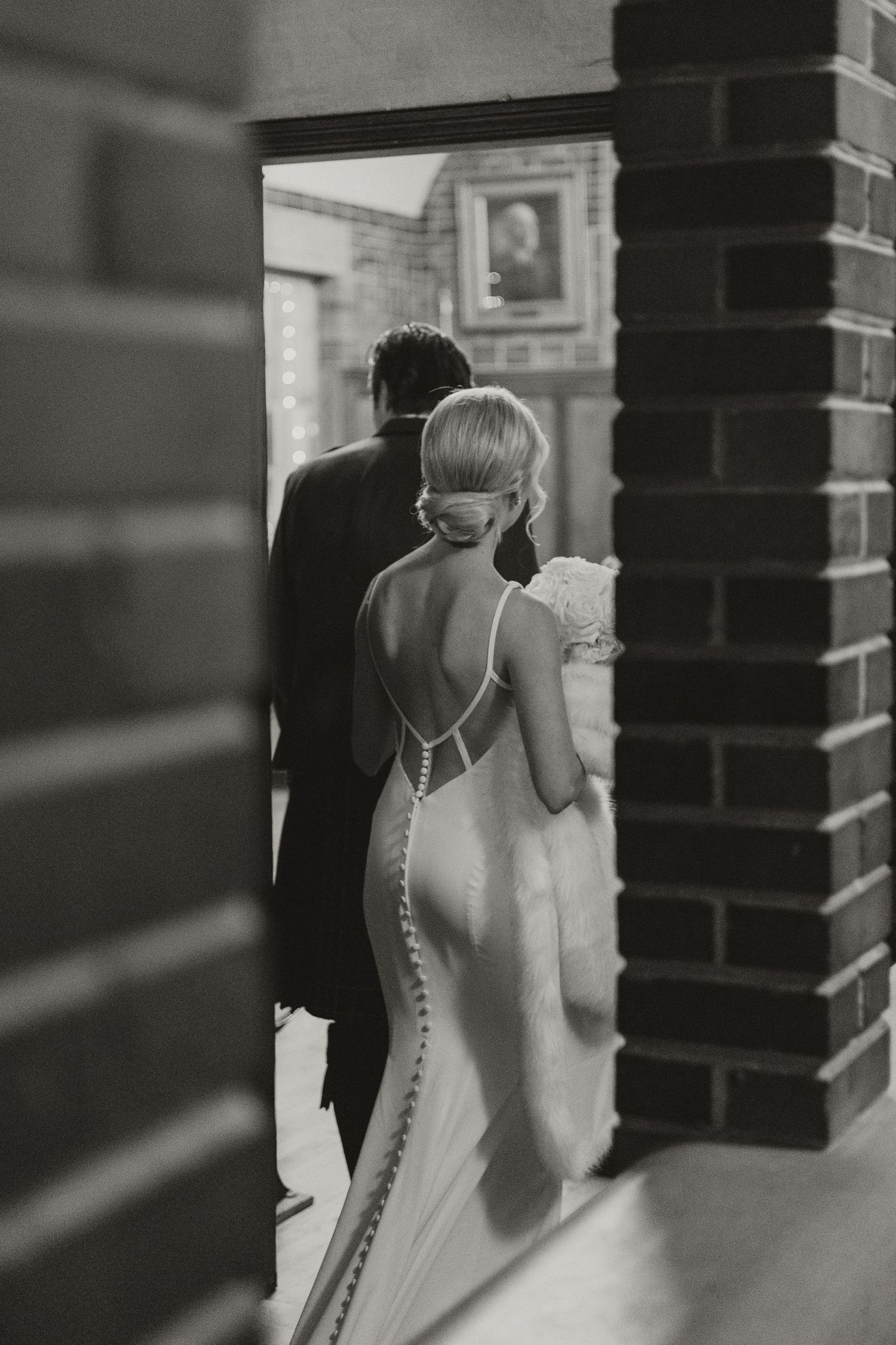 Black and white photo of bride in backless gown with button detail walking with groom through a brick archway.