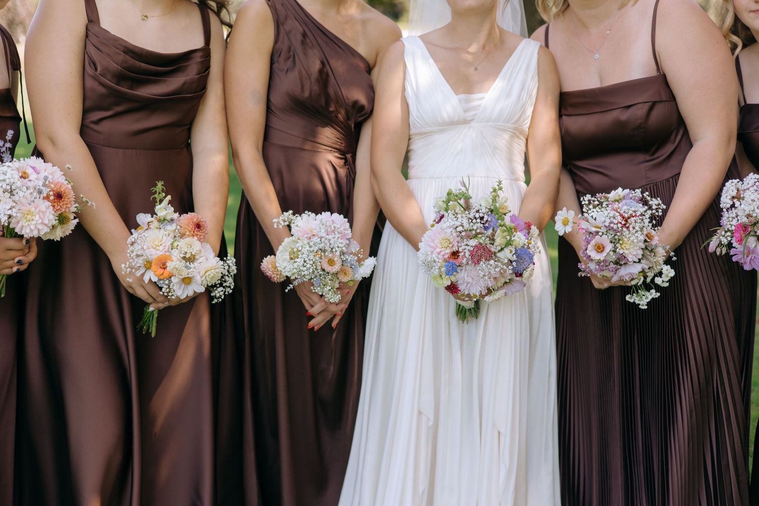 Bride in white gown with bridesmaids in brown satin dresses holding colorful floral bouquets at wedding.