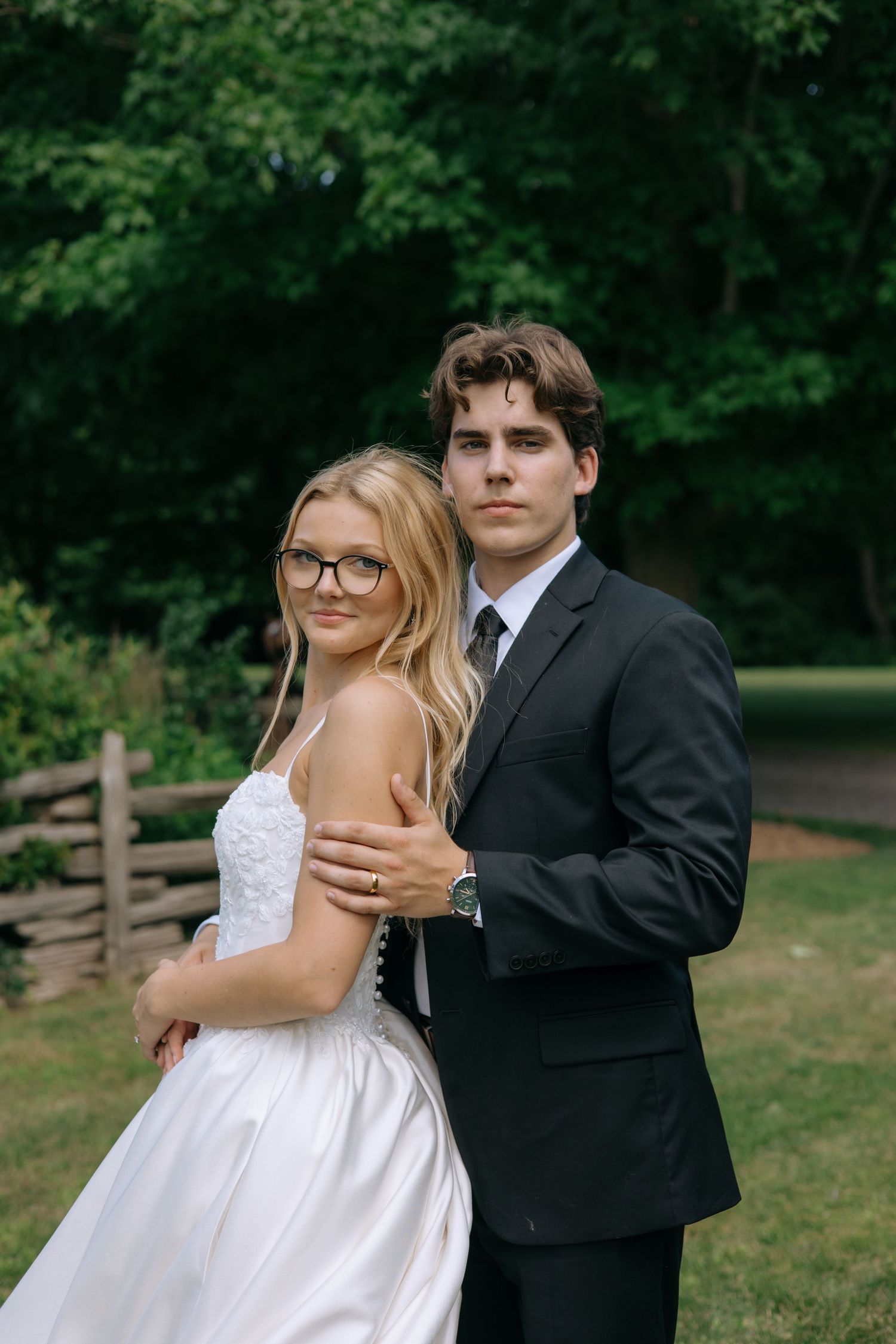 A bride in a white gown and groom in a black suit pose together outdoors near a wooden fence and lush green trees.