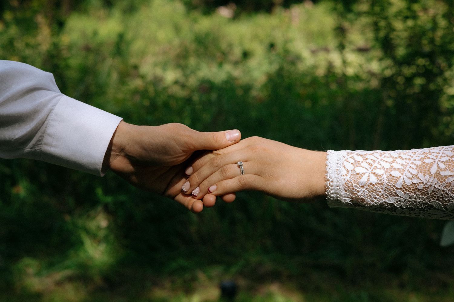 Couple holding hands outdoors, bride wearing lace sleeve and diamond engagement ring, groom in white dress shirt.