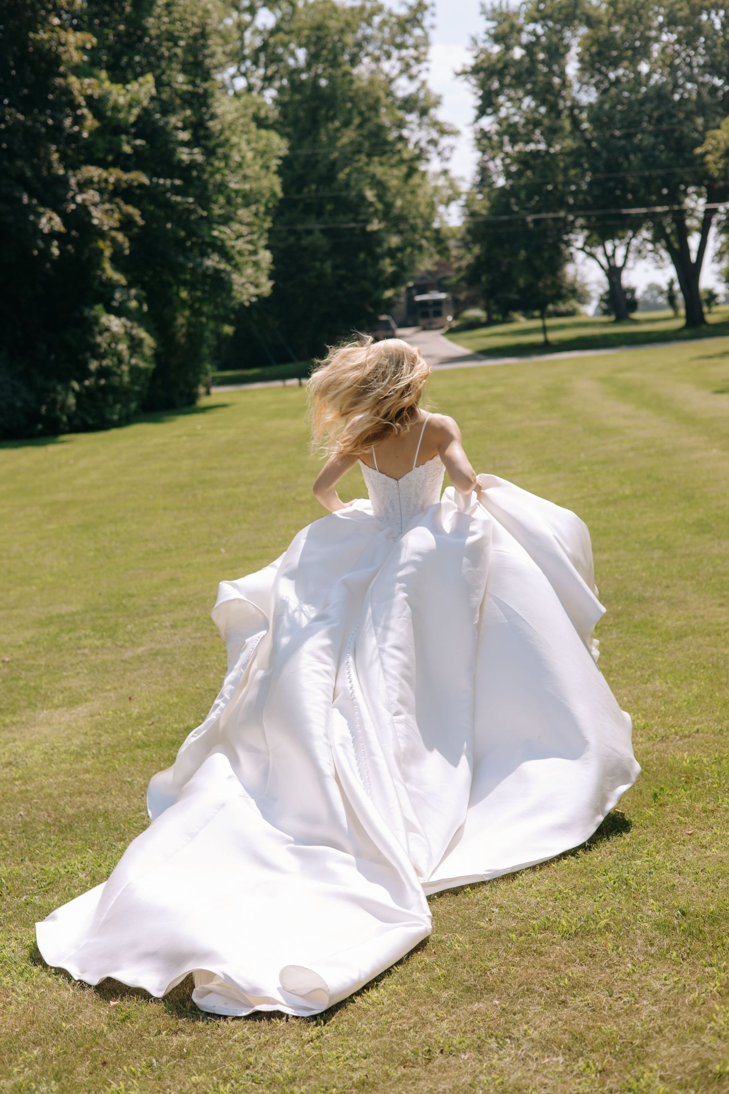 Bride in flowing white ball gown running across lush green lawn with hair blowing, back view on sunny day.