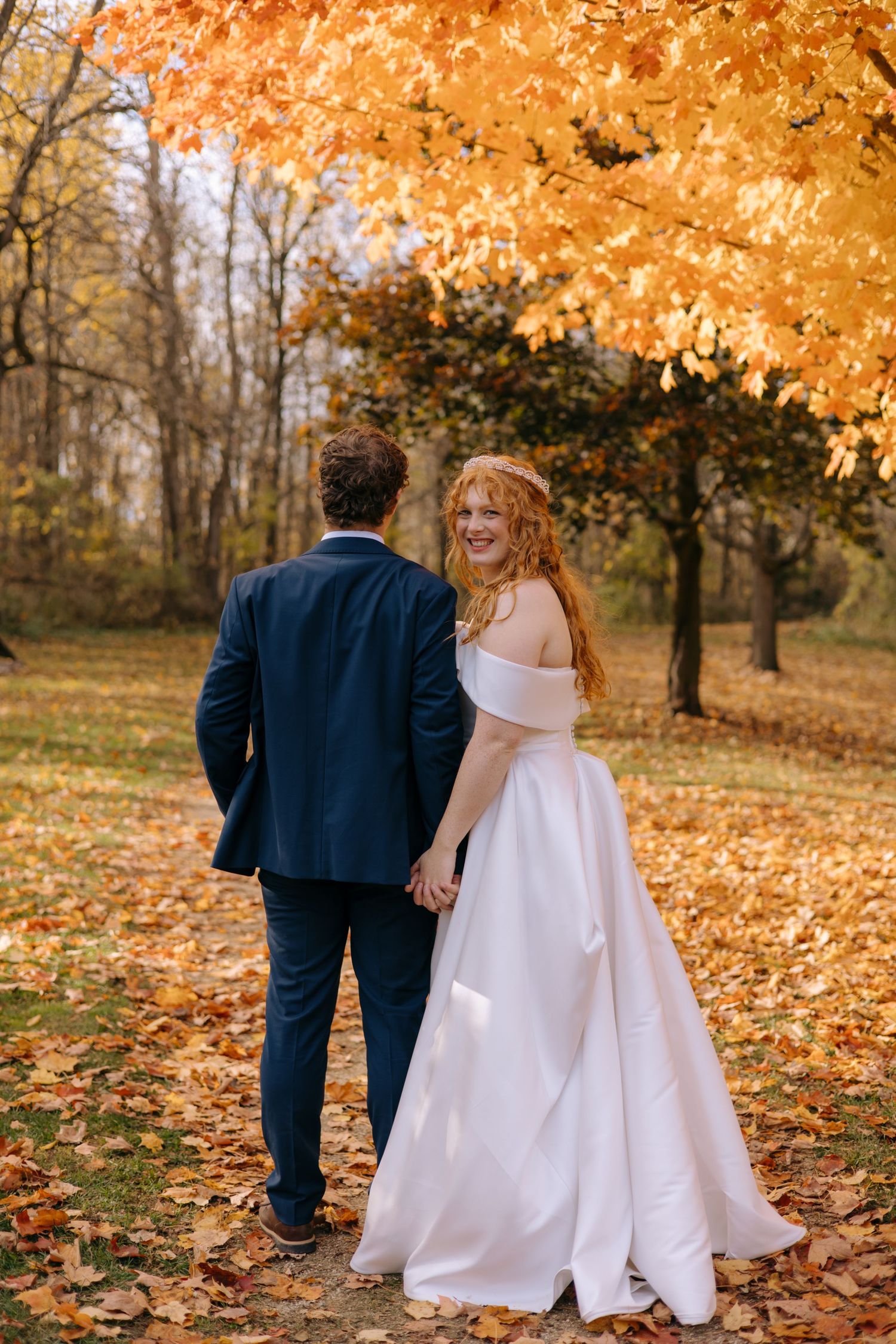 Bride in white off-shoulder gown looks back smiling while holding grooms hand in colorful autumn park.
