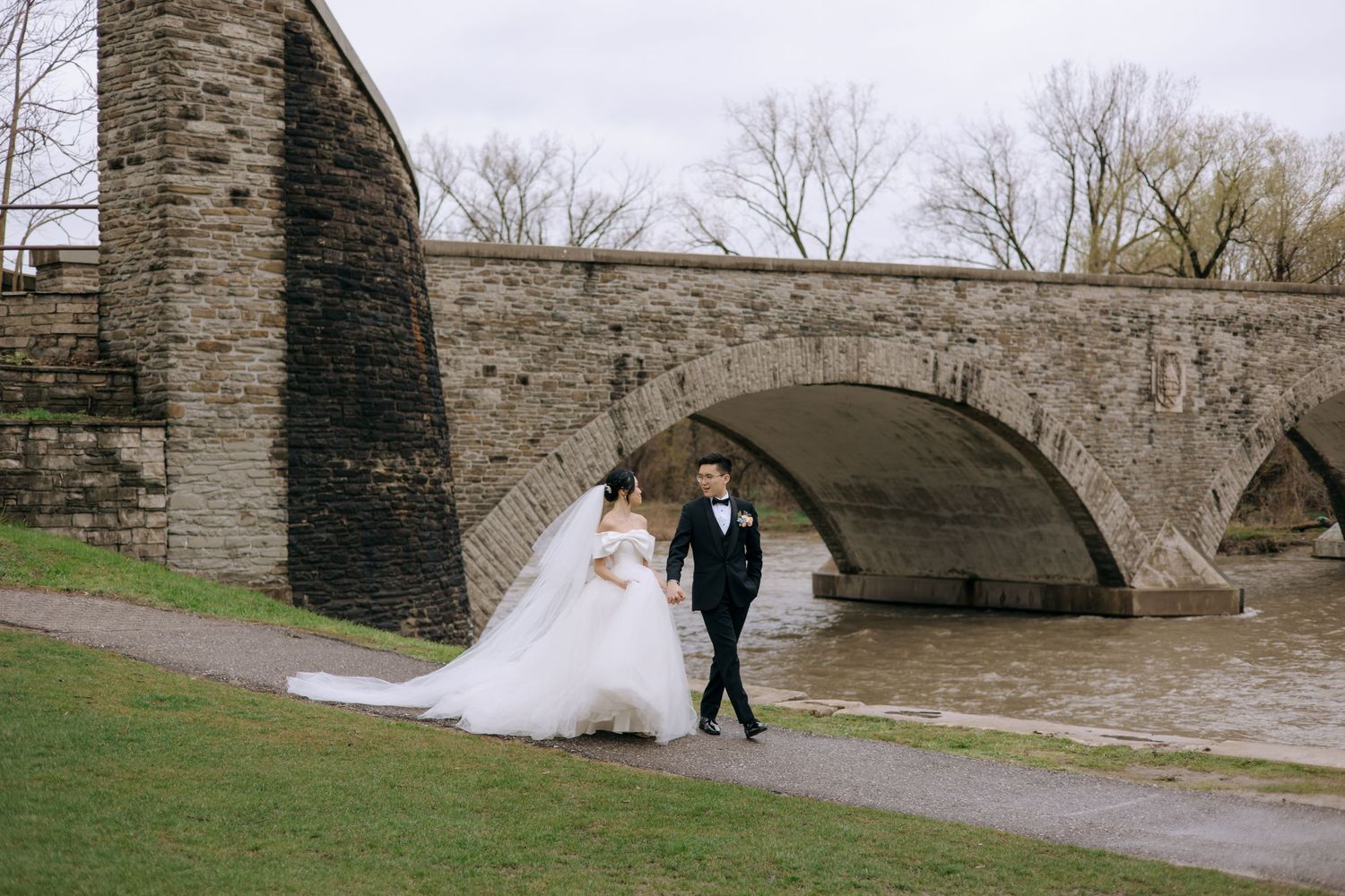 Bride and groom walking hand in hand along a river path beside a historic stone arch bridge.
