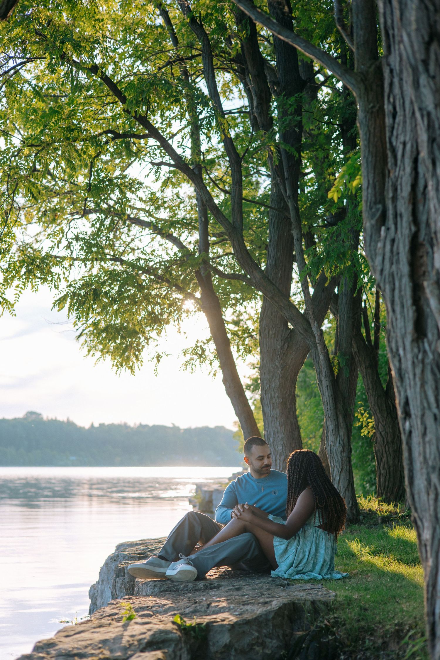 Couple sitting together on rocks by a serene river at golden hour, surrounded by lush green trees.