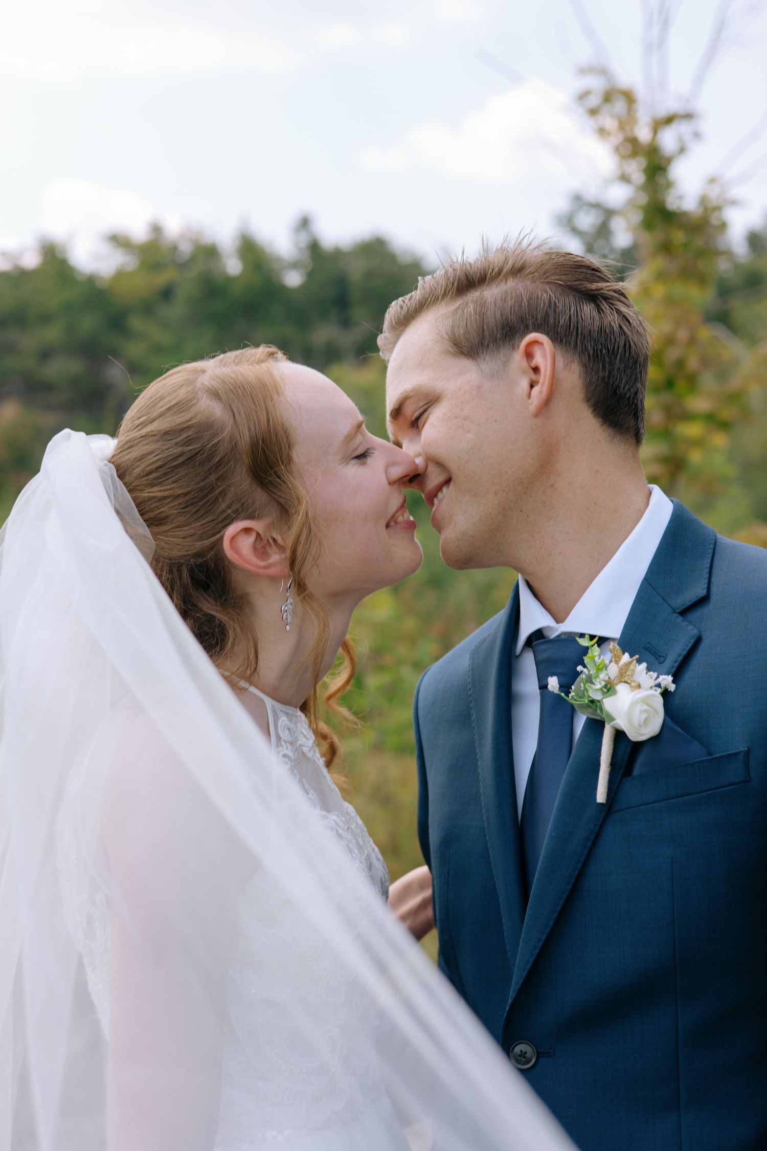 Bride and groom share a tender kiss outdoors, with the bride's veil flowing and groom wearing a navy suit with boutonniere.