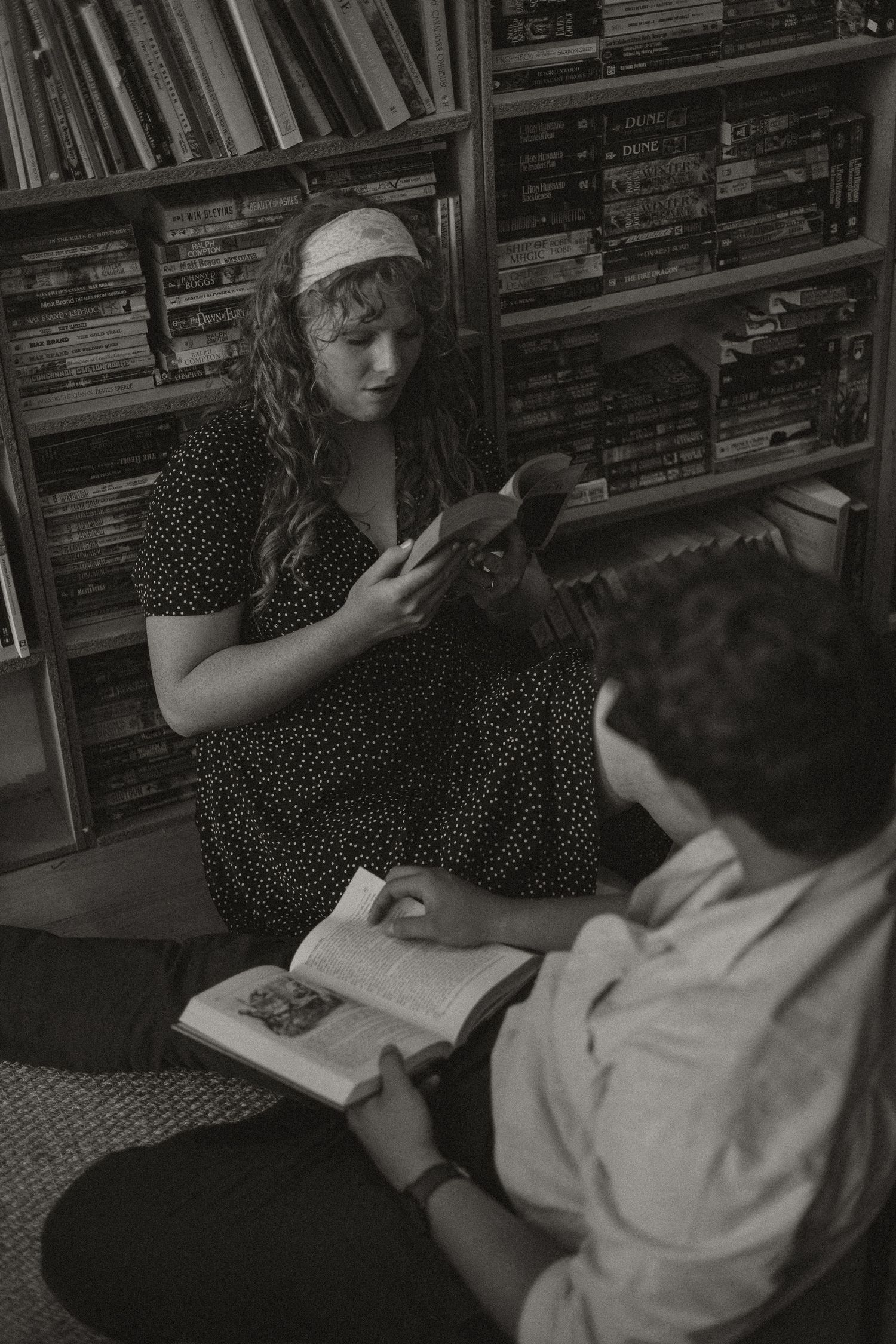 Black and white photo of a couple sitting on a floor reading books in front of a large bookshelf.