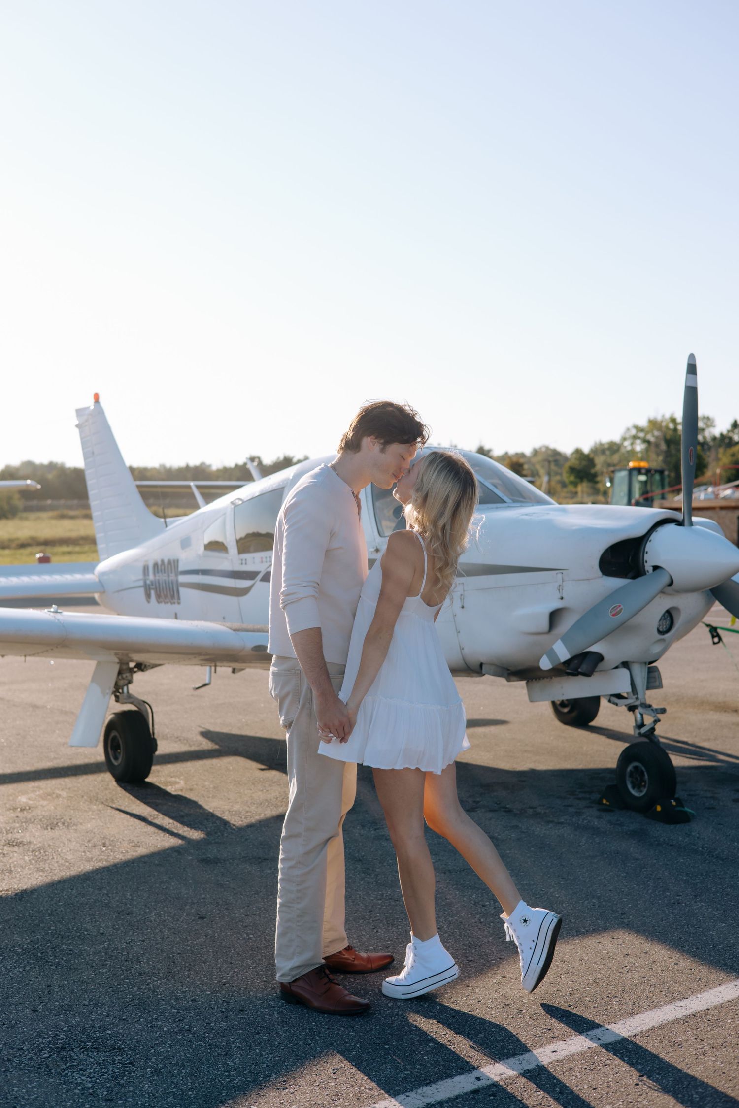 Romantic couple holding hands and nearly kissing in front of a small white propeller plane on a sunny airfield.