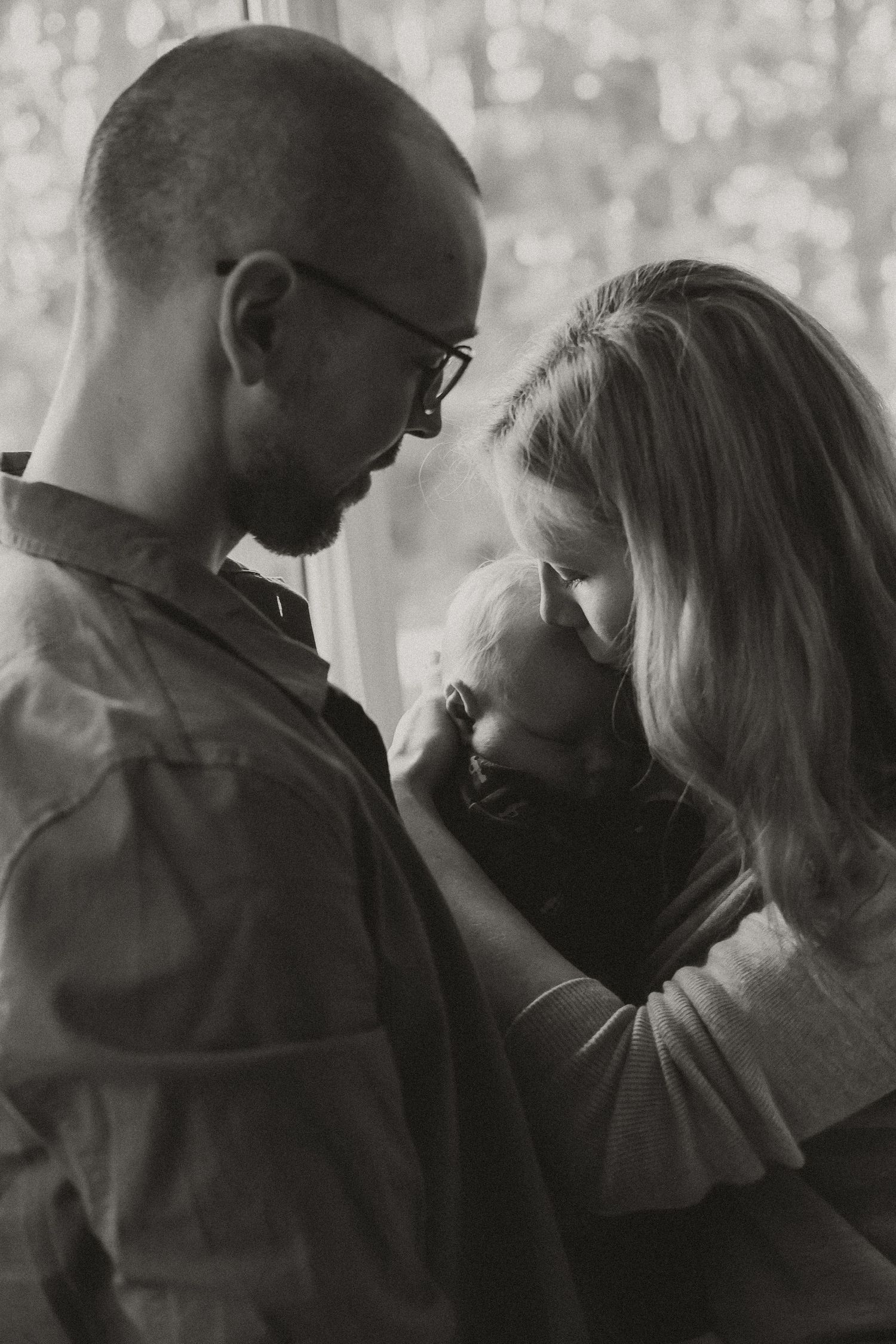 Black and white portrait of parents tenderly holding their newborn baby together by a window.