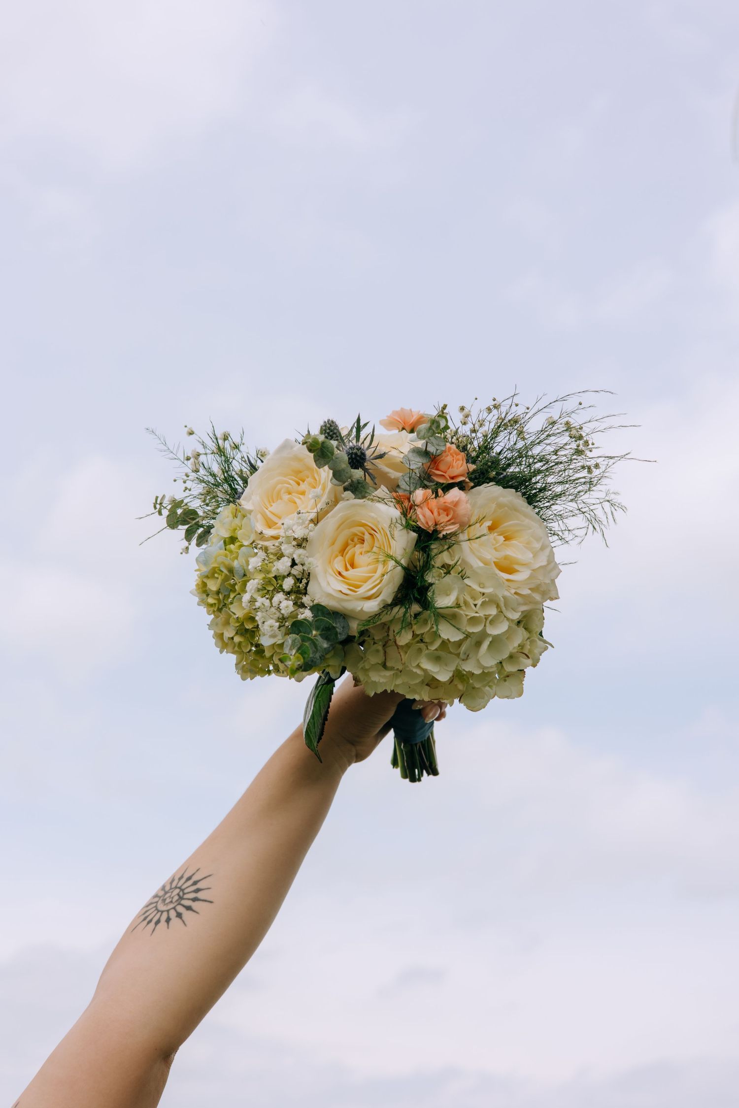 Hand with a sun tattoo holds up a bridal bouquet of white roses and hydrangeas against a cloudy sky.
